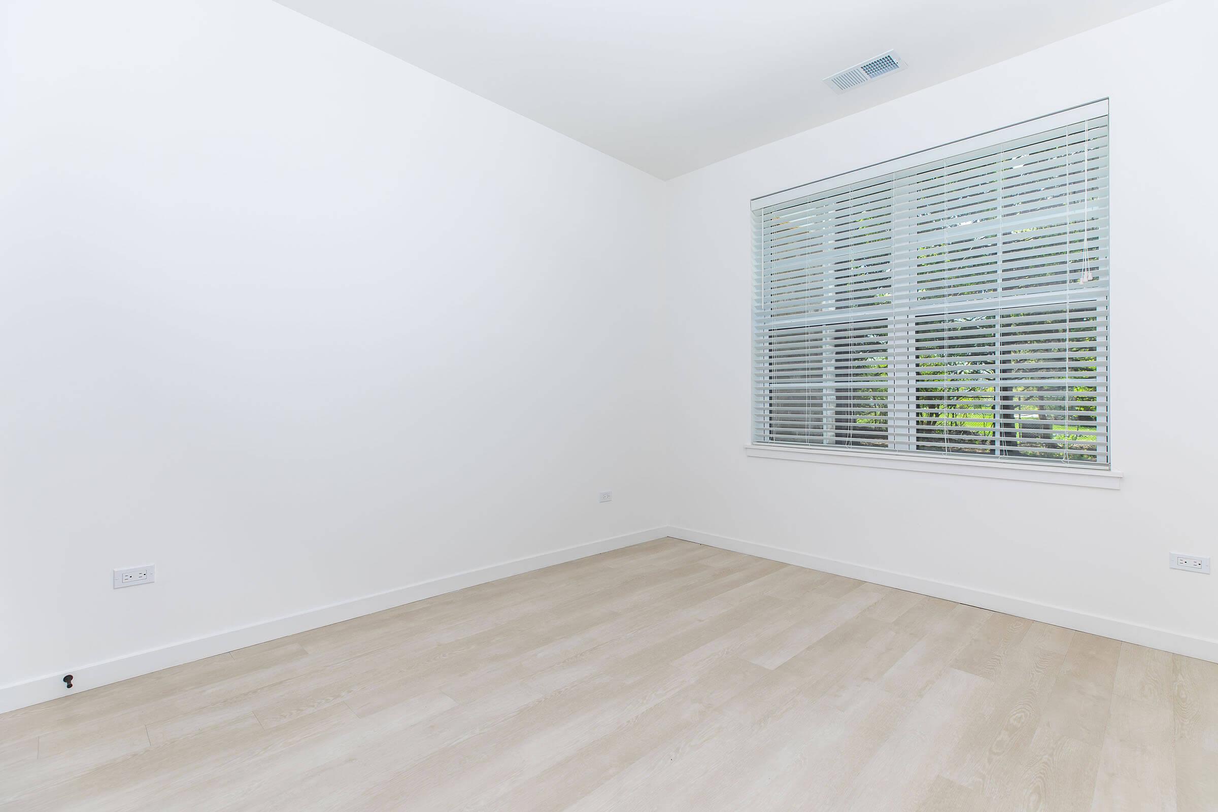 A spacious, empty room with white walls and light wood flooring. A large window with blinds allows natural light to enter, illuminating the space. The room has no furniture or decor, creating a minimalist and clean aesthetic.