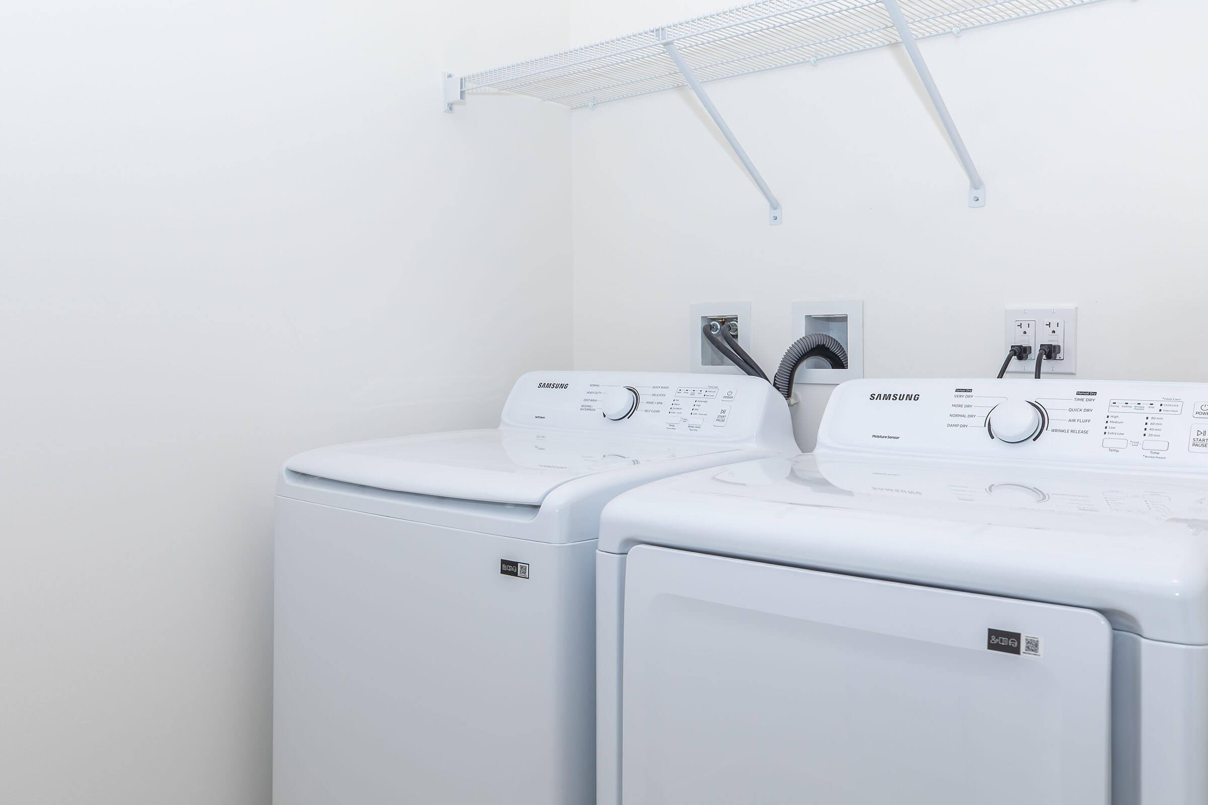 A clean laundry room featuring a white washing machine and dryer side by side. Above them, there is a wire shelf mounted on the wall for storage. The walls are plain and light-colored, creating a minimalist and tidy appearance.