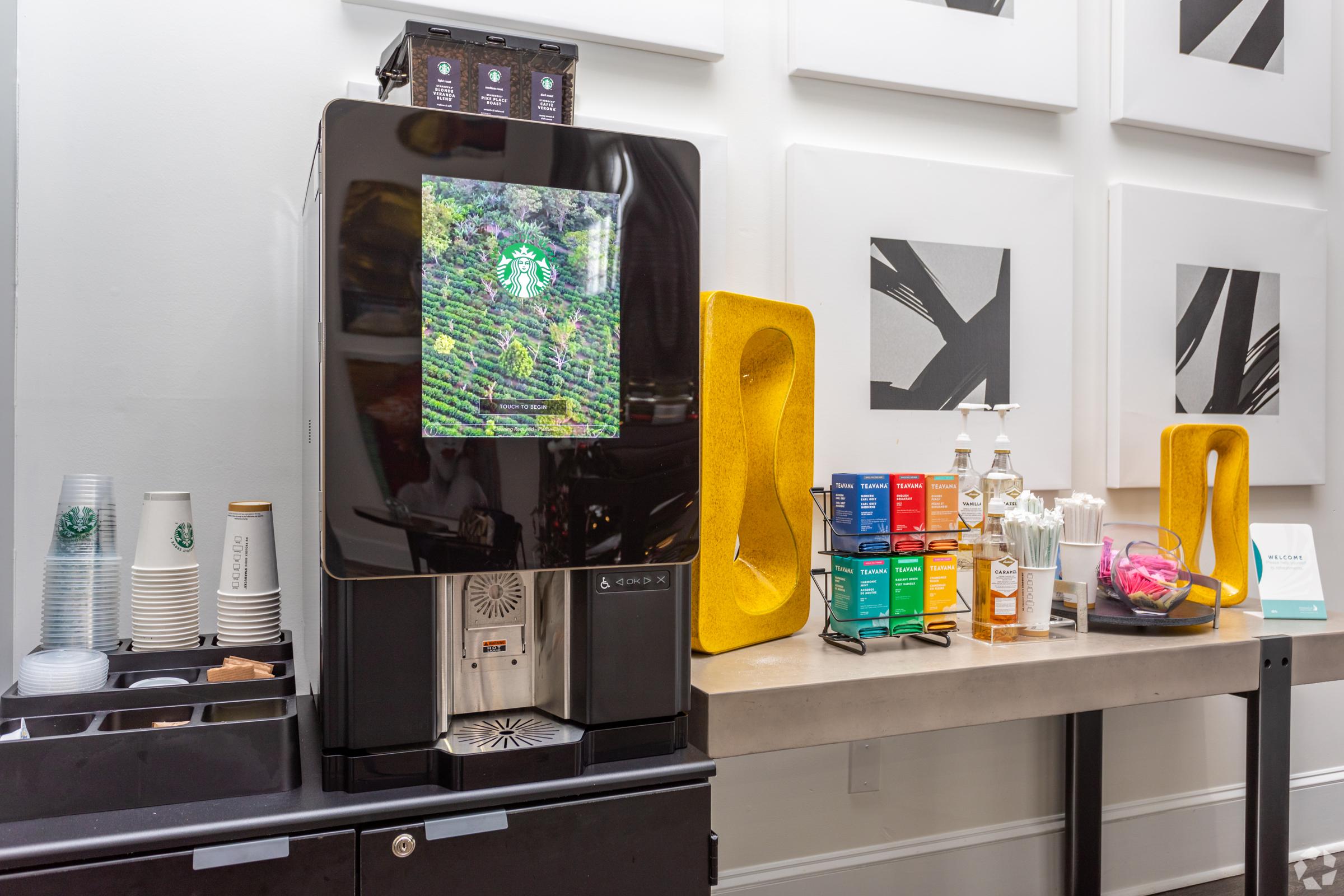 Coffee machine with a display showcasing coffee beans and a decorative setup featuring colored teas, sugar, and utensils. Nearby, there are cups and a small sign on a table against a backdrop of abstract artwork on the wall.