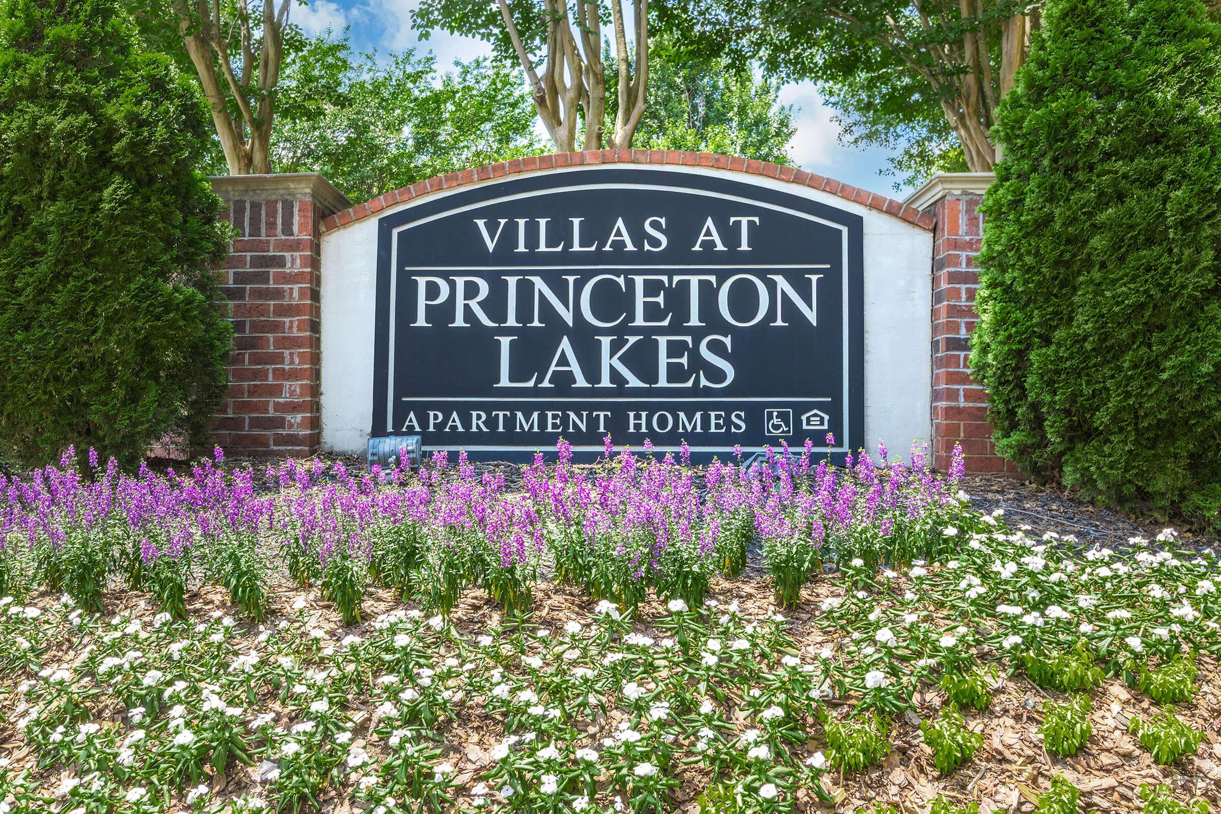 Sign for "Villas at Princeton Lakes" displaying the name prominently with a background of green bushes and colorful flowers in the foreground. The sign is set against a sunny blue sky, indicating a welcoming apartment community.