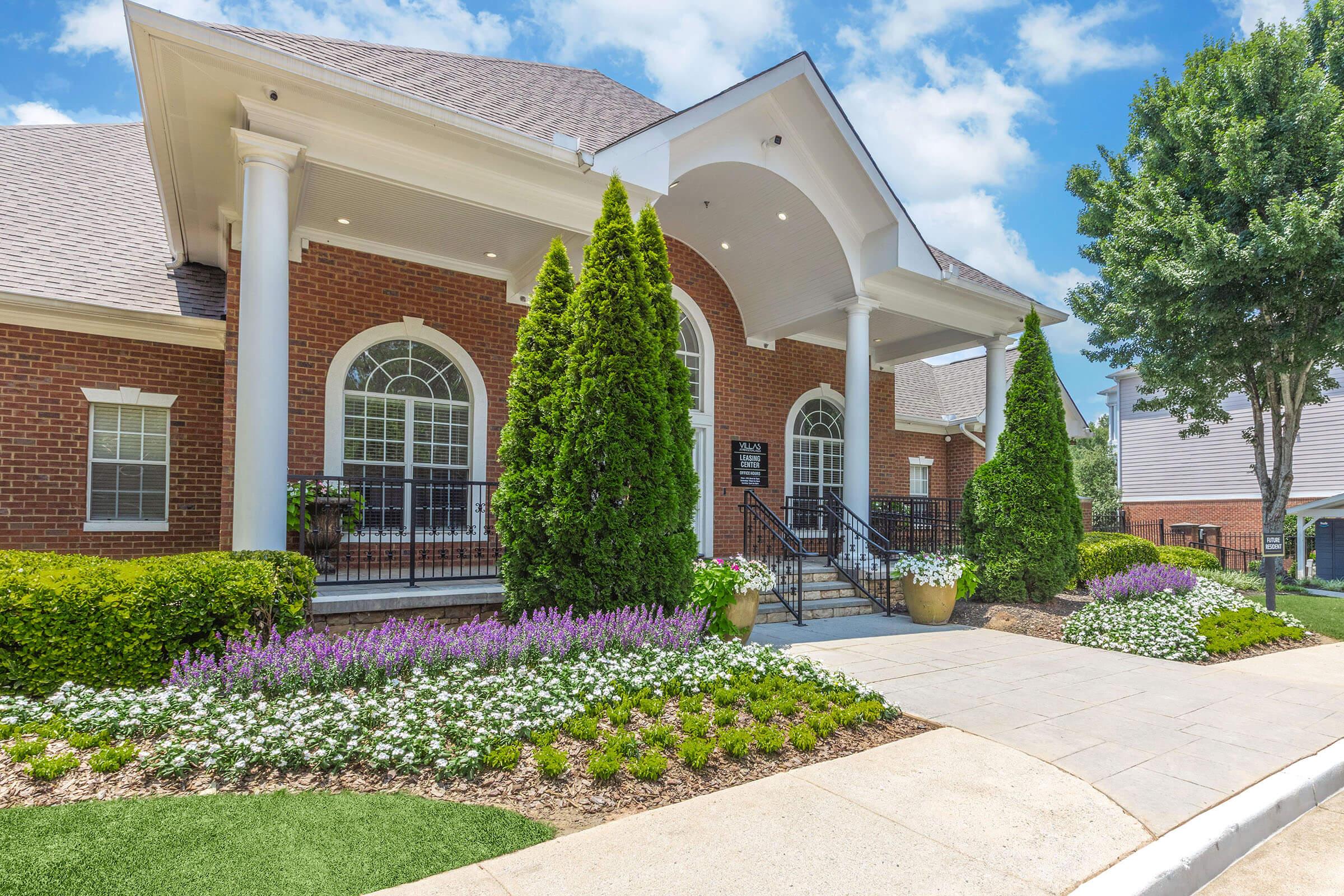 A modern brick building with a large entrance, featuring a white portico and decorative columns. Lush green landscaping includes manicured hedges, flowering plants in shades of purple and white, and tall coniferous trees. The walkway is paved with light-colored stones leading to the entrance.
