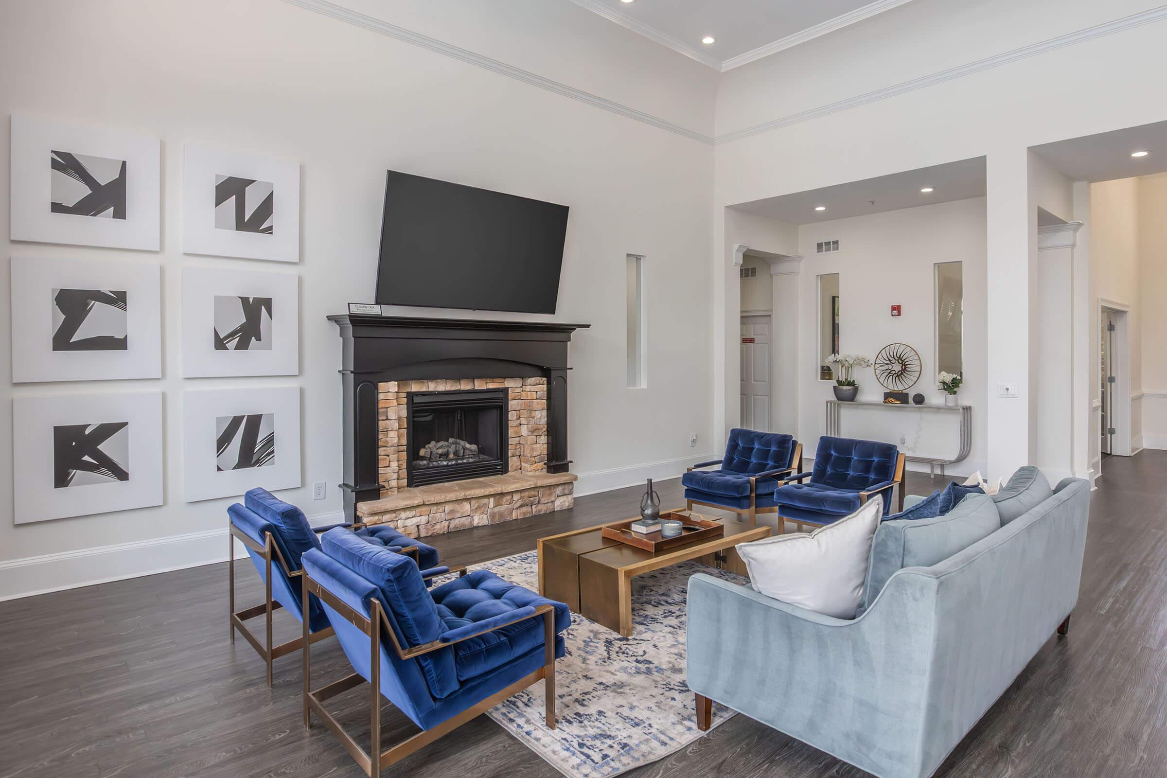 A modern living room featuring a cozy seating arrangement with blue velvet chairs and a light blue sofa. The space includes a fireplace with a brick surround, a large television mounted above it, and abstract art displayed on the walls. Stylish decor elements and natural light create a welcoming atmosphere.