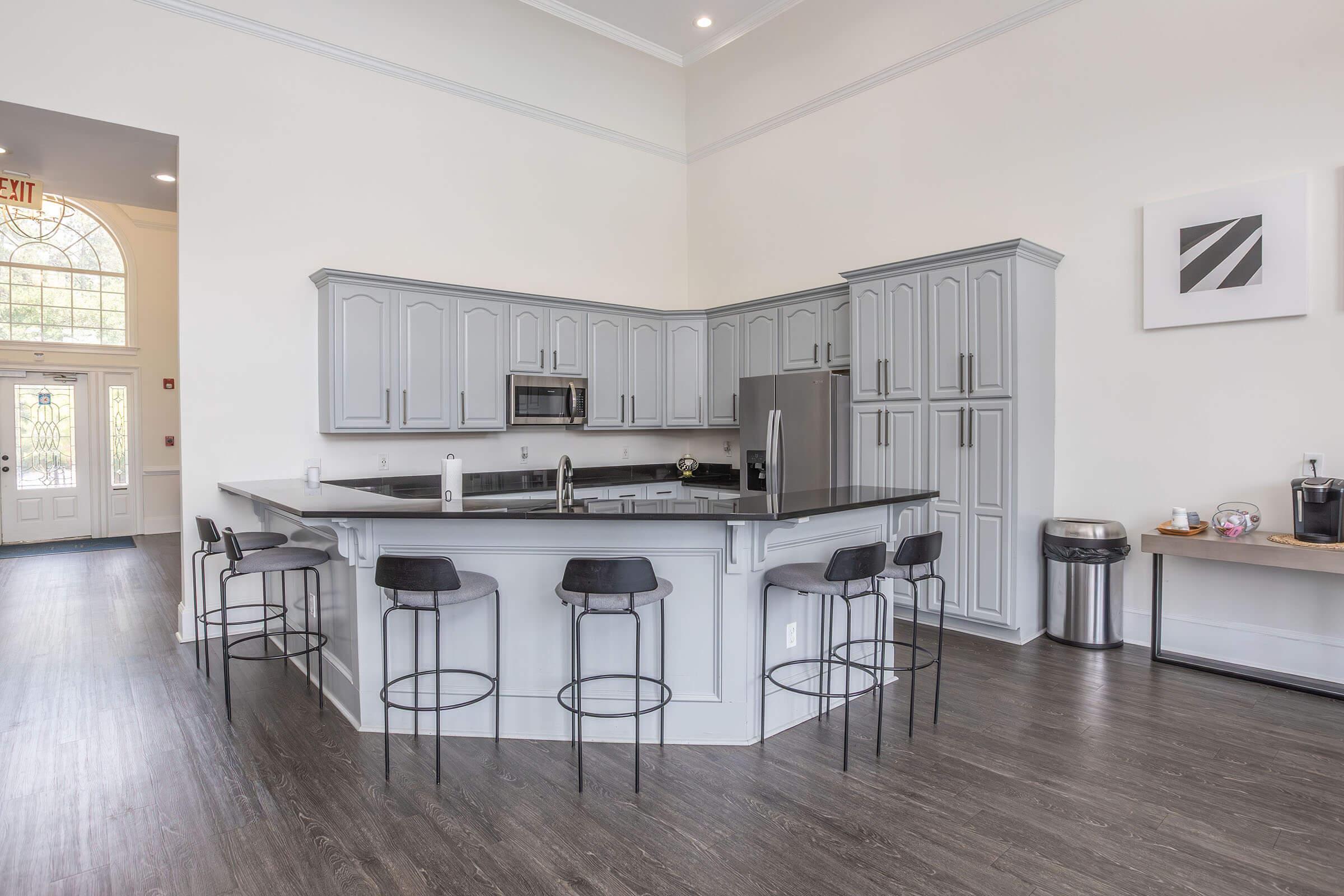 Modern kitchen interior with gray cabinetry and a large central island. The island features black countertop and seating for four on stylish stools. Appliances include a microwave and refrigerator, with a coffee machine on a side table. The space is bright and airy with light-colored walls and wooden flooring.