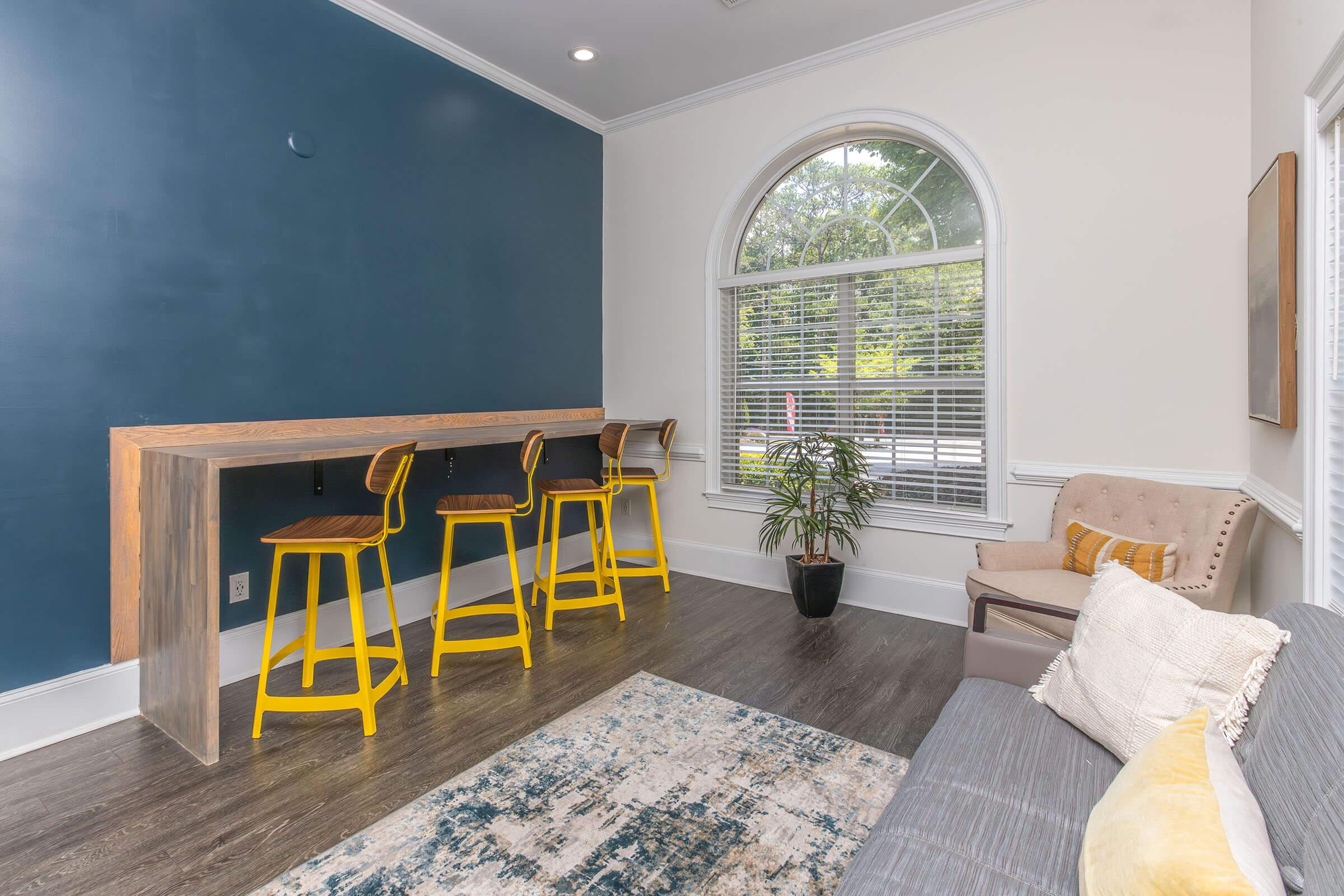 A cozy interior space featuring a wooden bar with three yellow stools, a beige couch, and a potted plant. Natural light streams through a large arched window, illuminating the room with a blue accent wall and a patterned area rug on the floor.