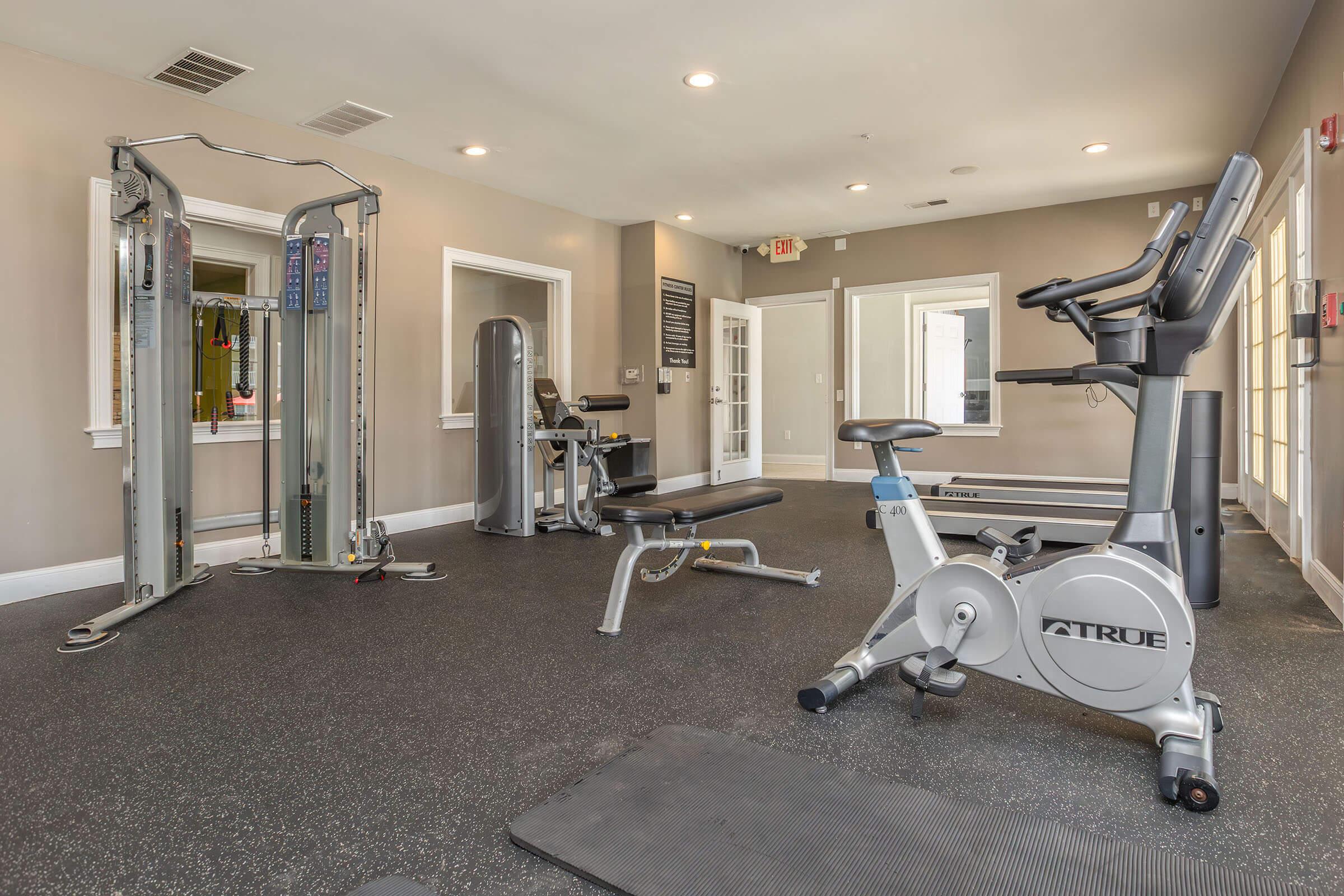 A modern gym interior featuring various workout equipment, including a stationary bike, weight machine, and benches. The space has a sleek design with gray walls and a rubber floor, creating an inviting atmosphere for fitness activities. Natural light streams in through large windows.