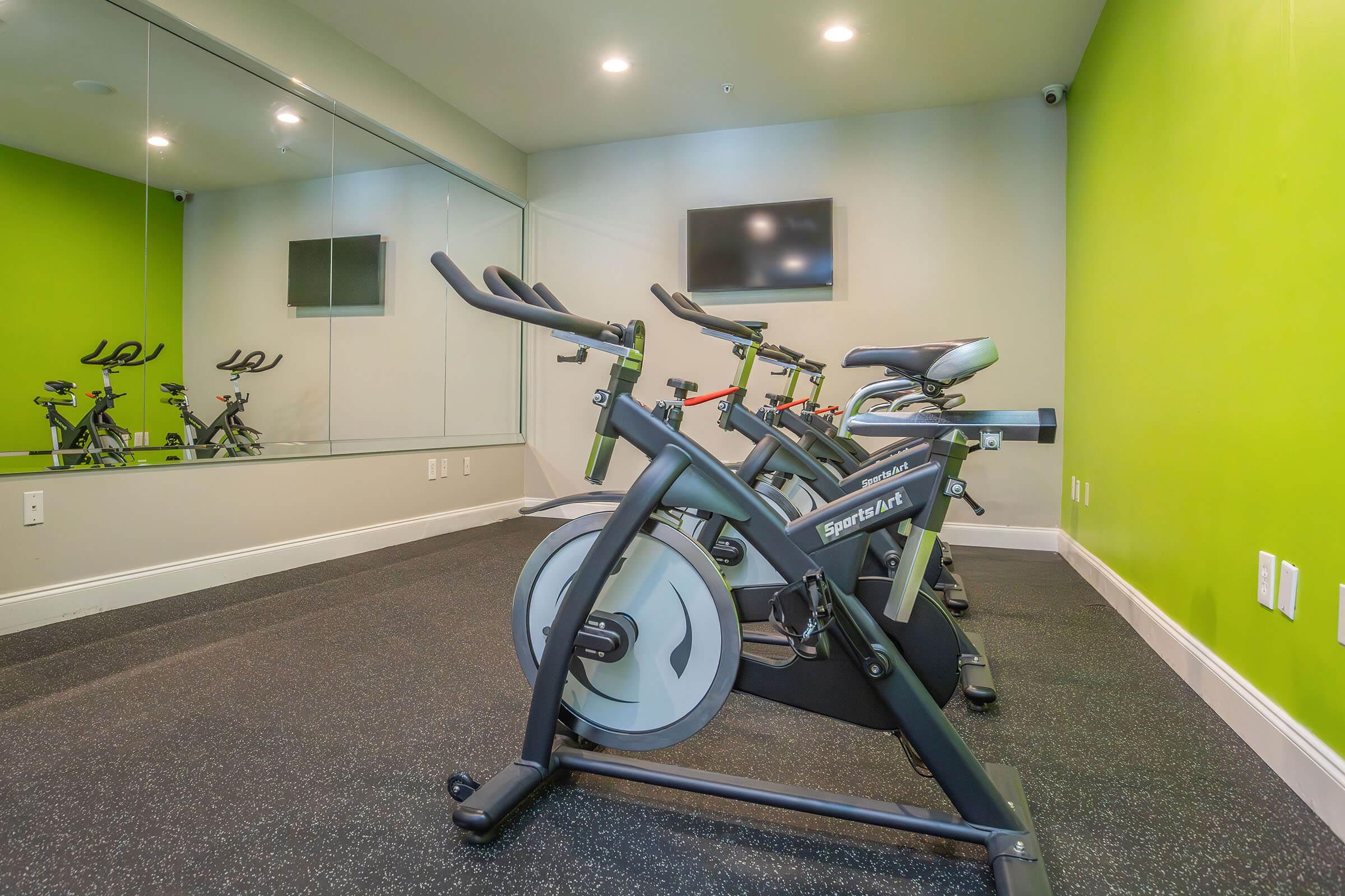 A brightly colored fitness room featuring several stationary bikes positioned on a black rubber floor. There is a large mirror on one wall and a television screen mounted on another, enhancing the modern workout atmosphere. The walls are painted a vibrant green, creating an energetic environment.