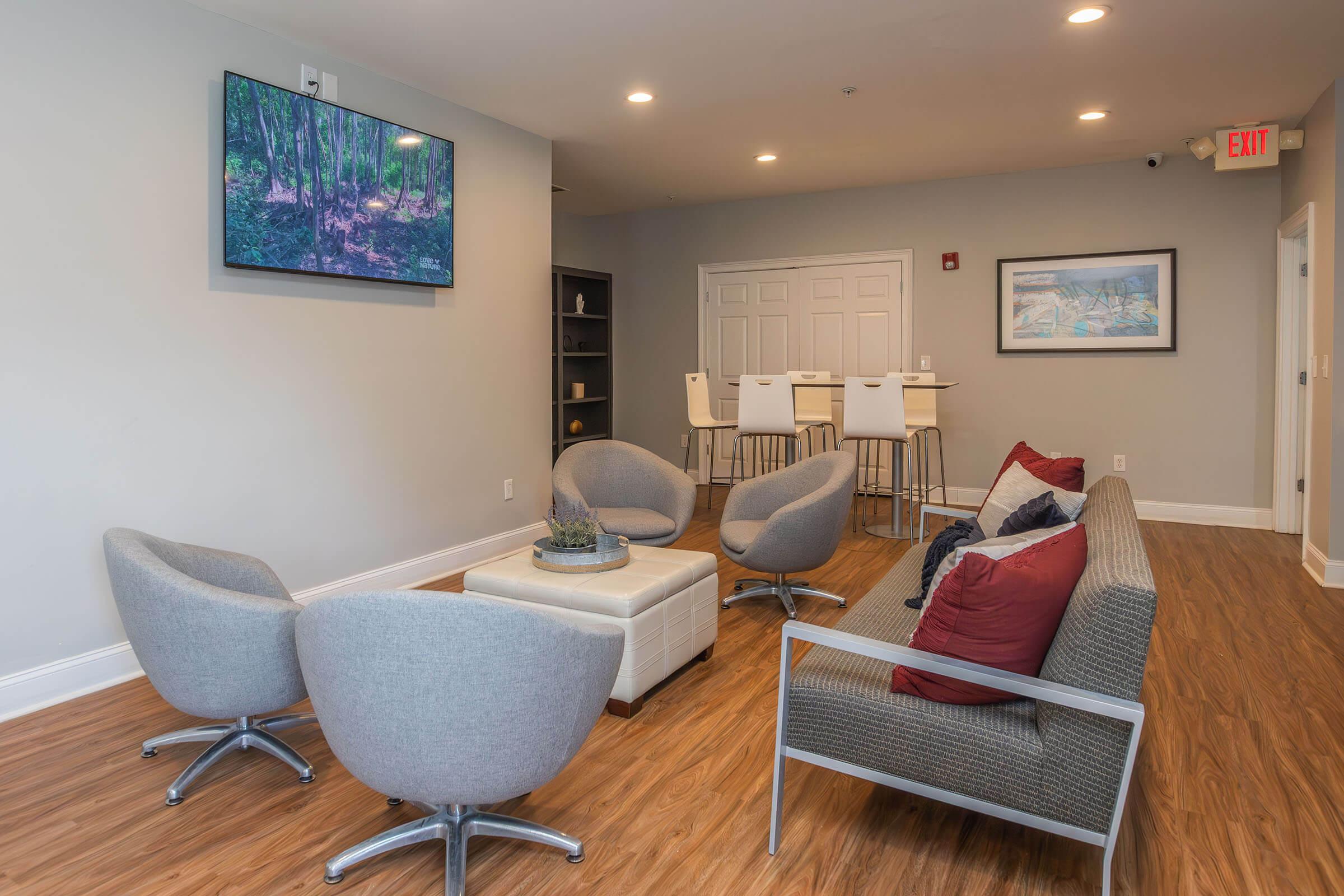 A modern indoor lounge area featuring four gray armchairs, a light-colored coffee table, and a sofa with red and gray cushions. A flat-screen TV displays a nature scene on the wall. In the background, there is a bar-style table with four high chairs and a shelf displaying decor. The flooring is hardwood.