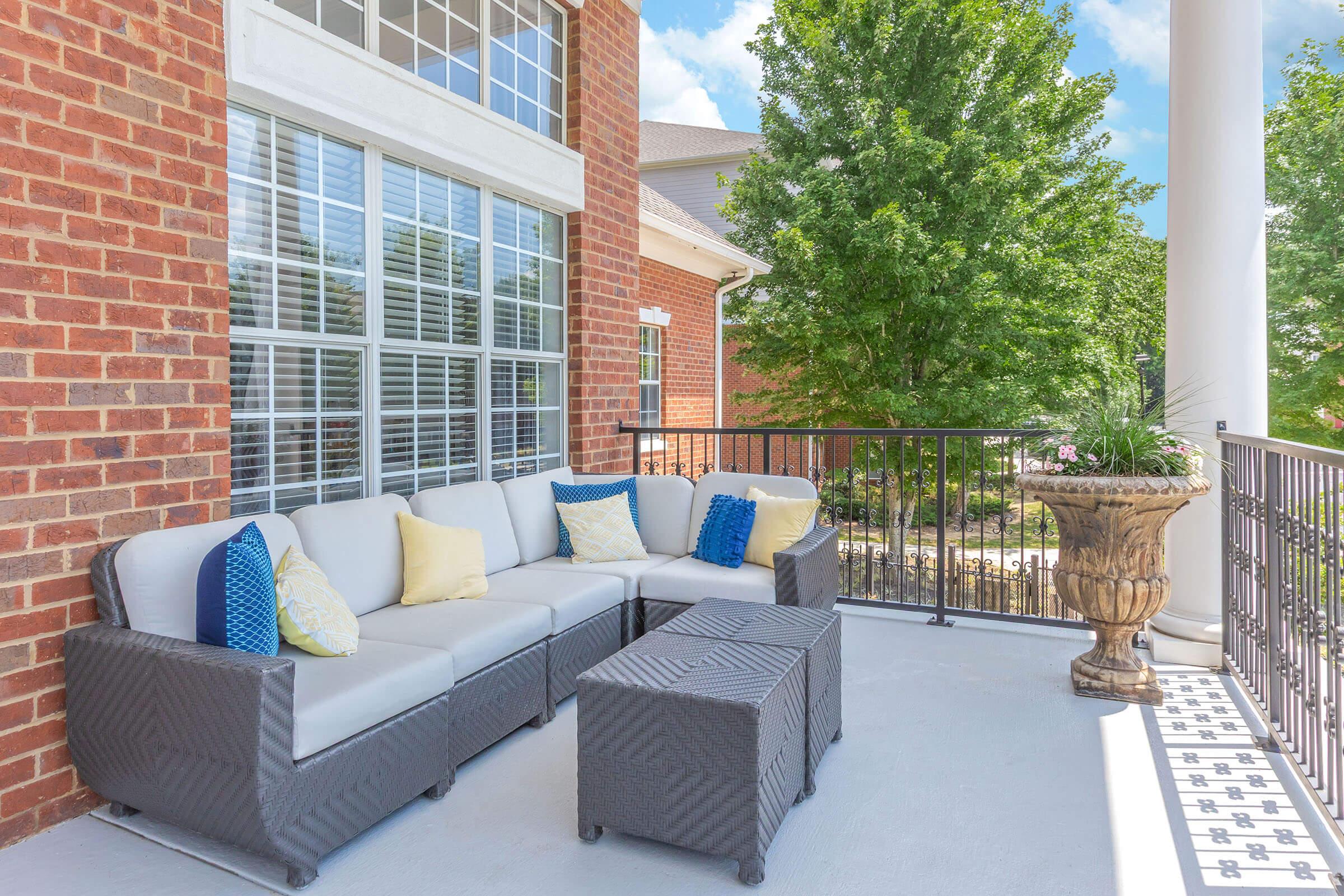 A cozy outdoor patio space featuring a light gray sectional sofa with yellow and blue throw pillows, accompanied by a small dark wicker table. The area has a lush green backdrop with trees and a decorative planter, illuminated by bright, natural light. Elegant brick walls add to the charm of the setting.