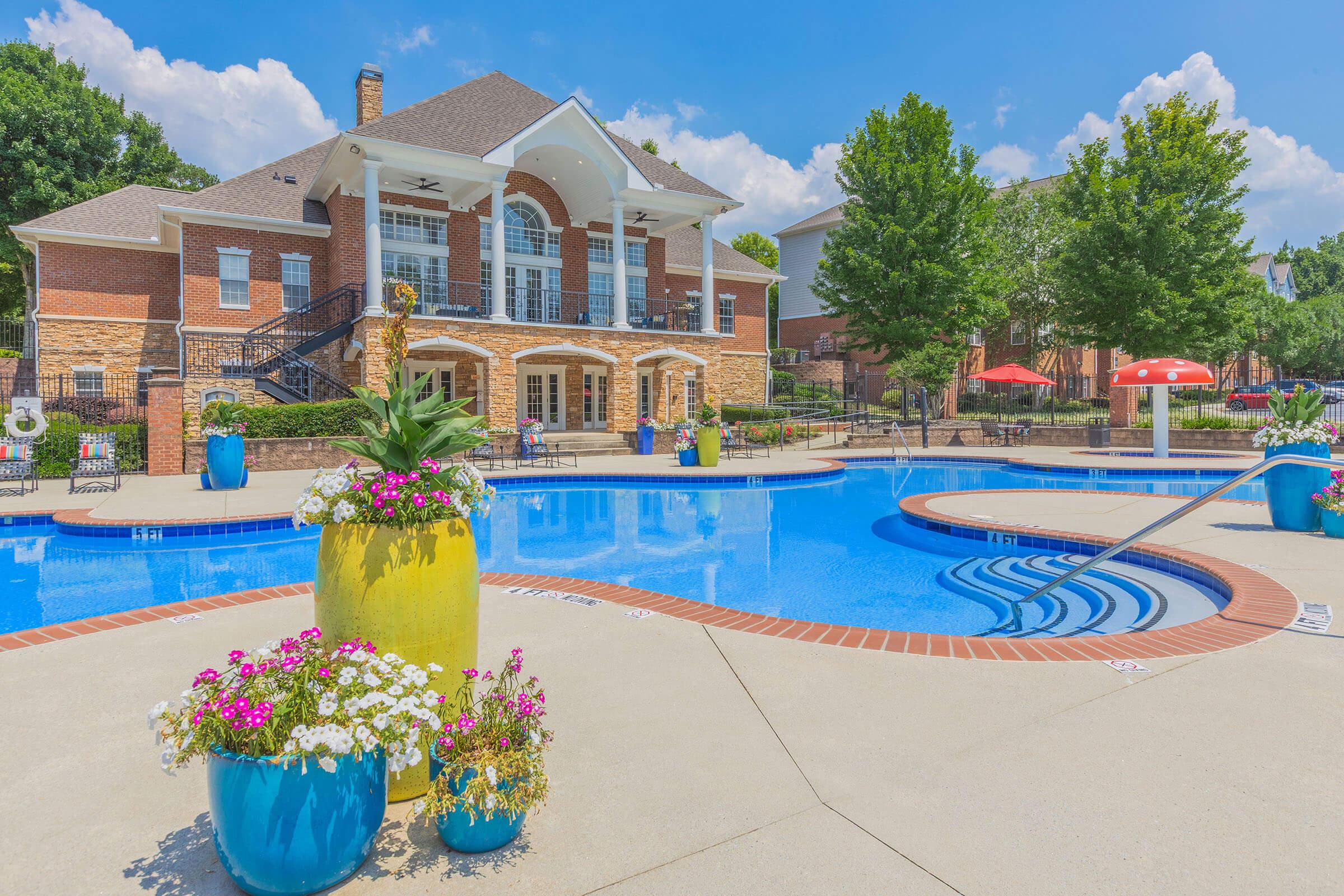A bright and inviting outdoor pool area featuring a clear blue pool surrounded by colorful flower pots. The pool is set against a spacious brick building with large windows. Lounge chairs and umbrellas provide shaded seating, and lush green trees can be seen in the background under a blue sky with fluffy white clouds.