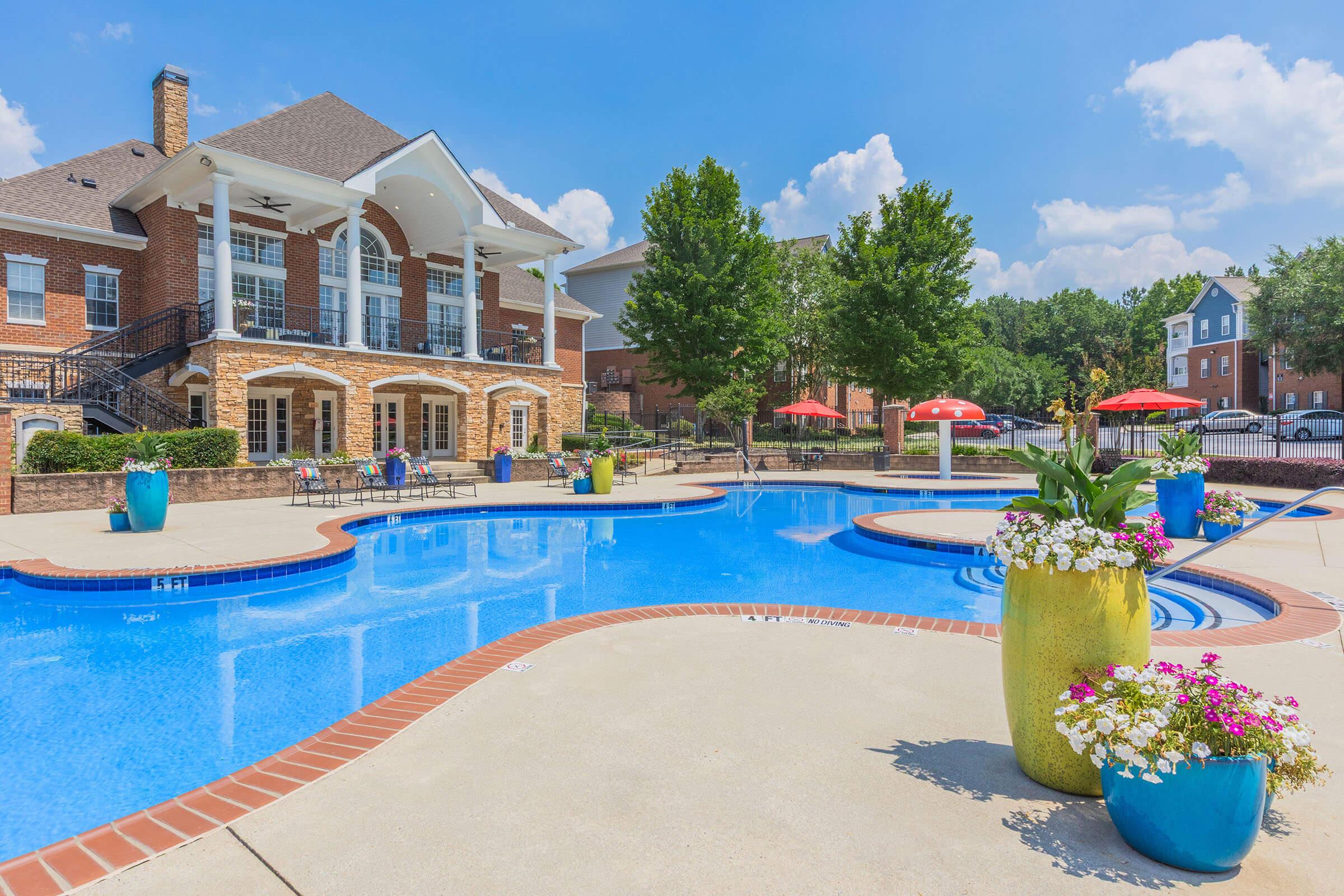 A luxurious outdoor pool area featuring a bright blue swimming pool surrounded by lounge chairs, vibrant planters with colorful flowers, and red umbrellas. In the background, there is a spacious brick building with a covered patio. The scene is set against a clear blue sky with fluffy white clouds.