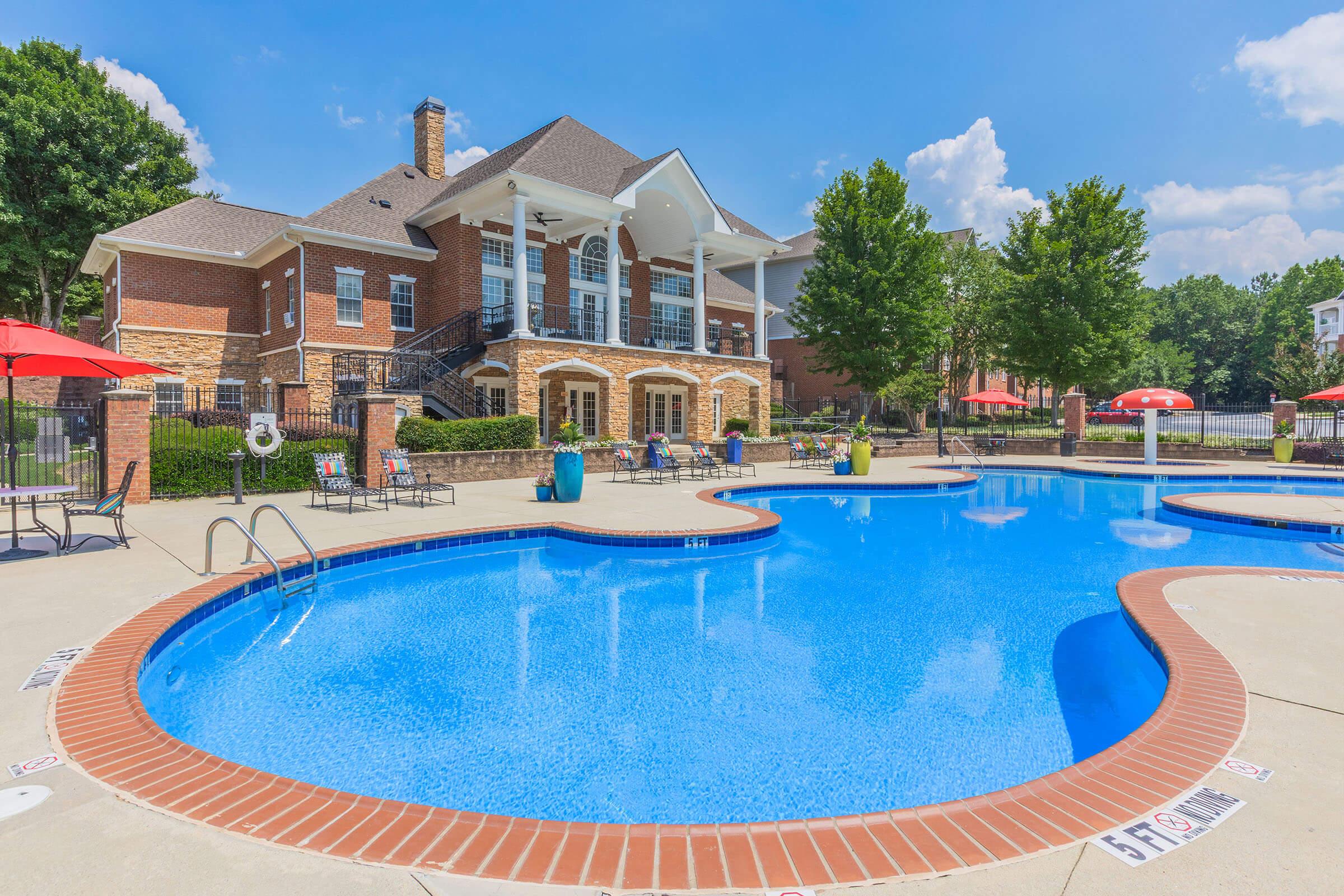 A bright blue swimming pool with a curved edge, surrounded by lounge chairs and umbrellas, in front of a large brick building with white columns. Lush green trees and a clear blue sky can be seen in the background, creating a serene outdoor atmosphere.