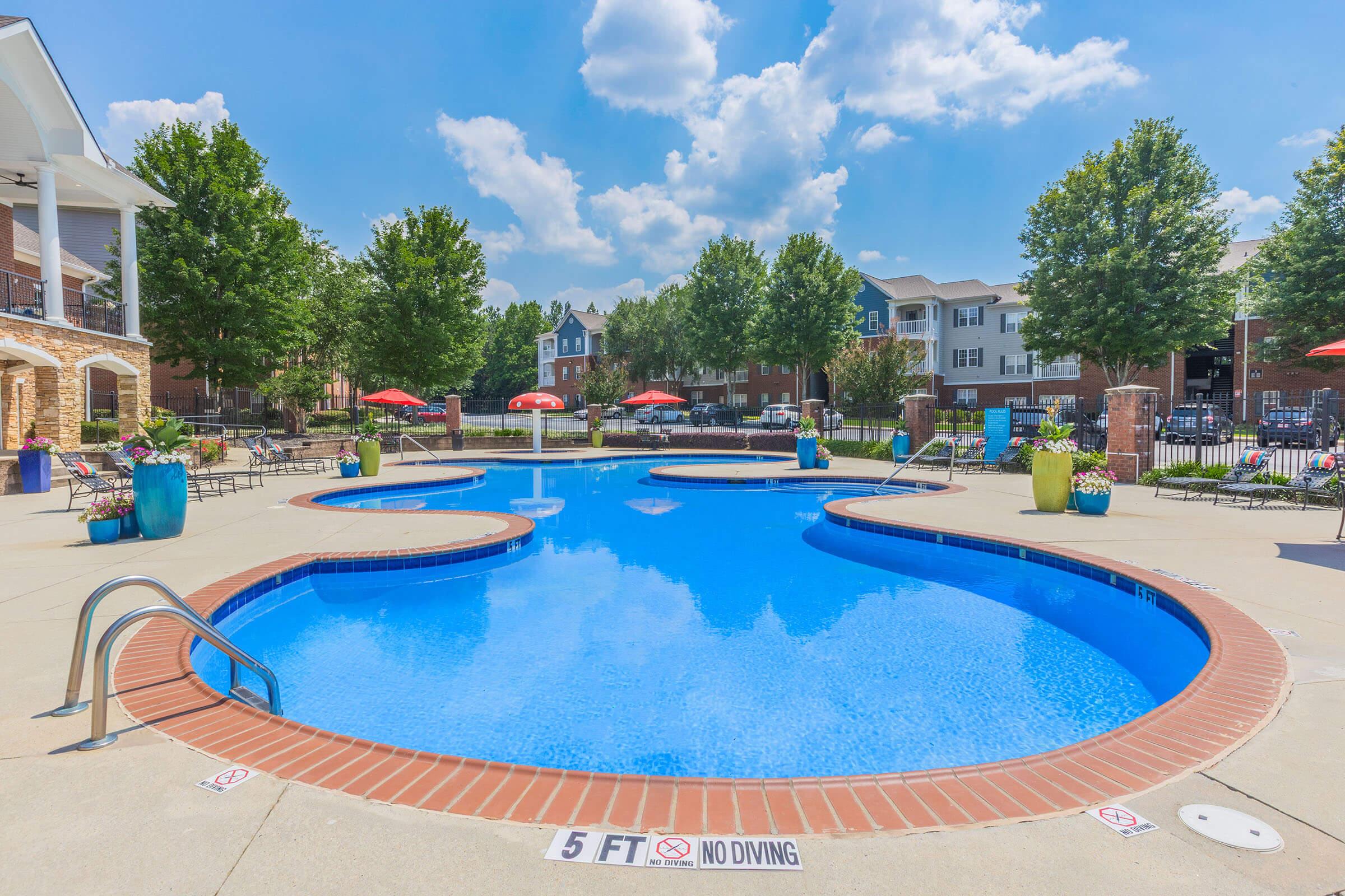 A bright, sunlit pool area featuring a curved swimming pool surrounded by lush green trees and lounge chairs. Red umbrellas offer shade, and colorful planters decorate the surroundings. A clear blue sky with fluffy white clouds completes the inviting atmosphere, with signage indicating the maximum depth of the pool.