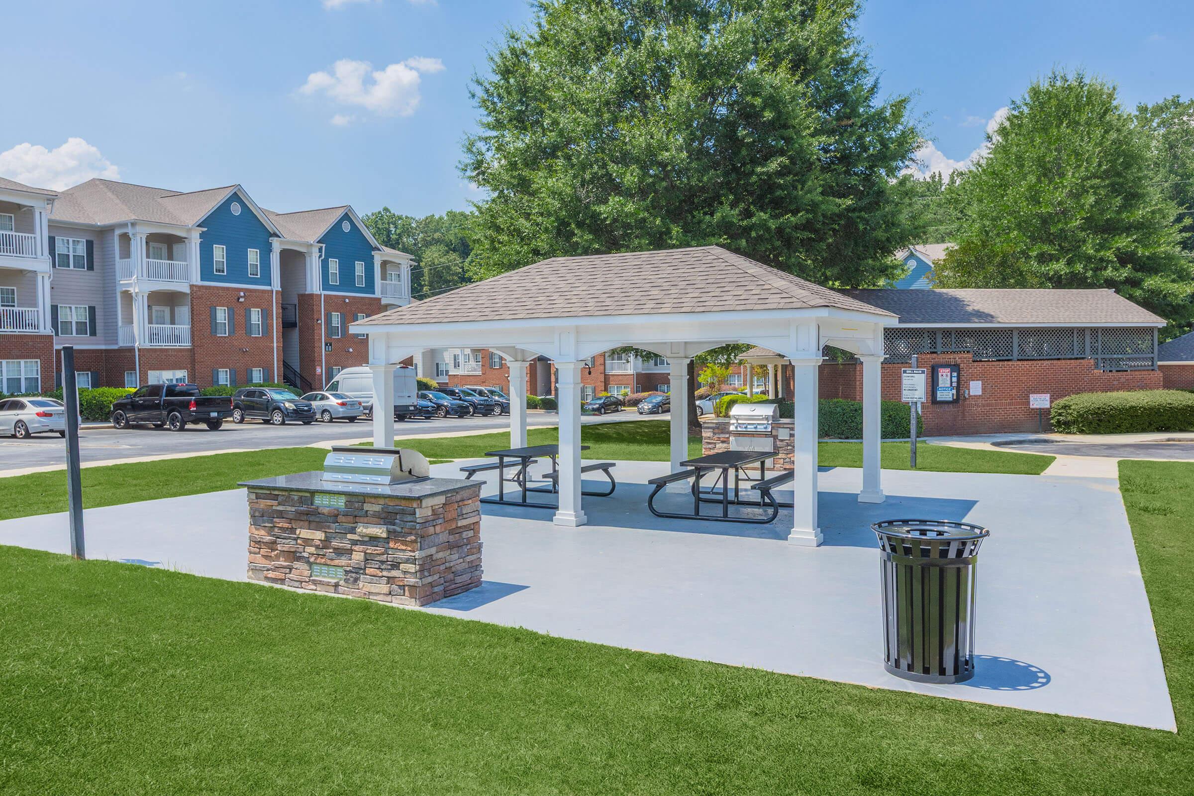 A spacious outdoor picnic area featuring a white gazebo with picnic tables and grill stations. Surrounding the area are well-manicured lawns and nearby apartment buildings, along with a trash can for convenience. Clear blue skies above create a bright, inviting atmosphere.