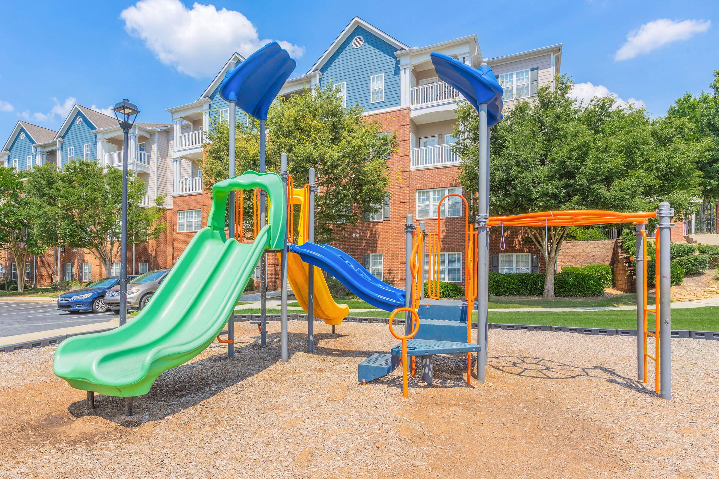 A colorful children's playground featuring a green slide and a blue slide, surrounded by orange structures. The playground is situated on gravel and is flanked by trees and apartment buildings in the background under a clear blue sky.