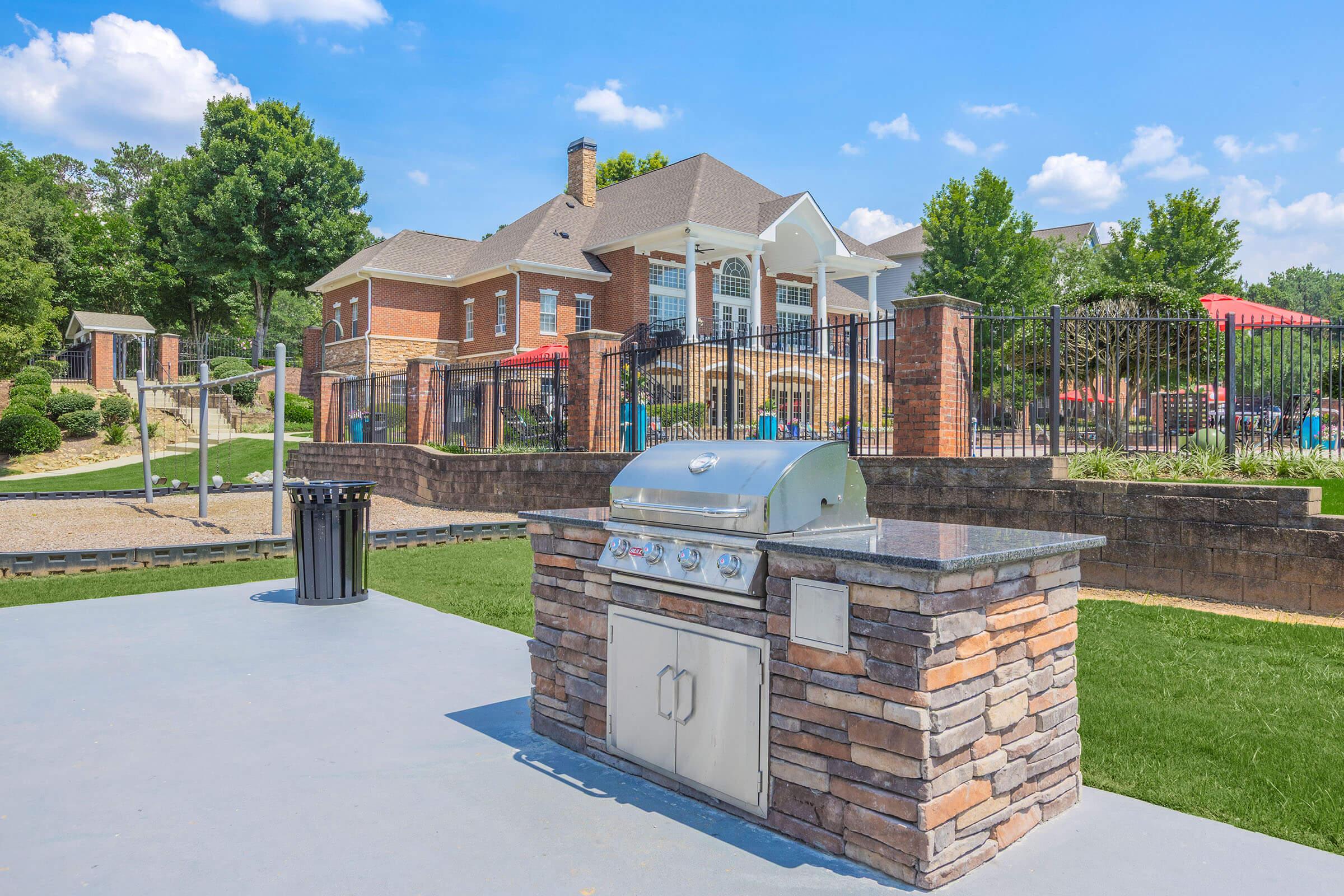Outdoor grill feature made of stone with a stainless steel surface, set in a landscaped area. In the background, a large brick building with multiple stories and a fenced yard is visible, surrounded by trees and recreational areas. The sky is bright blue with a few clouds.