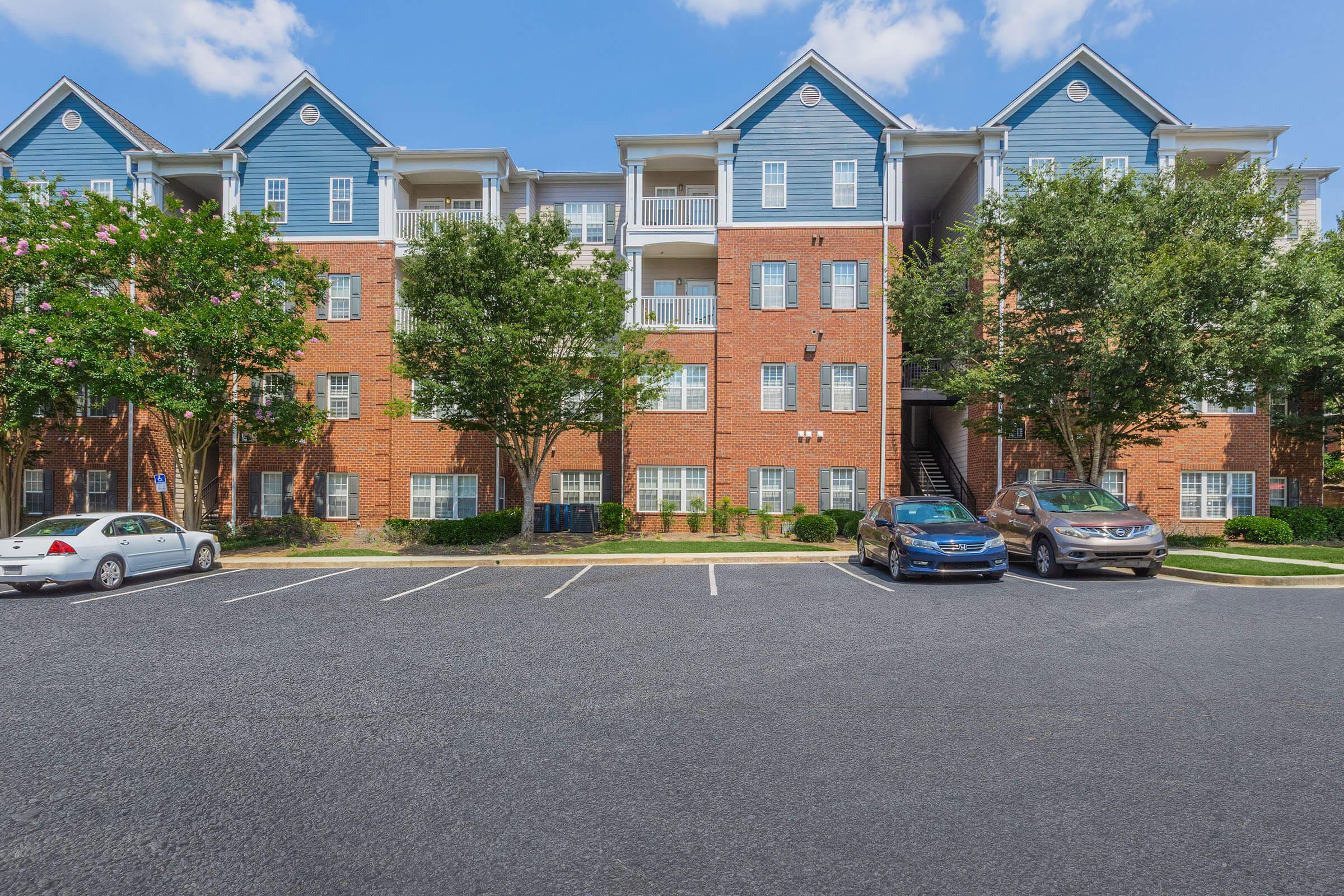 A multi-story brick apartment building with blue accents. The building features balconies and several windows, surrounded by trees and well-kept landscaping. A parking lot in the foreground has a few parked cars, with clear blue skies above.
