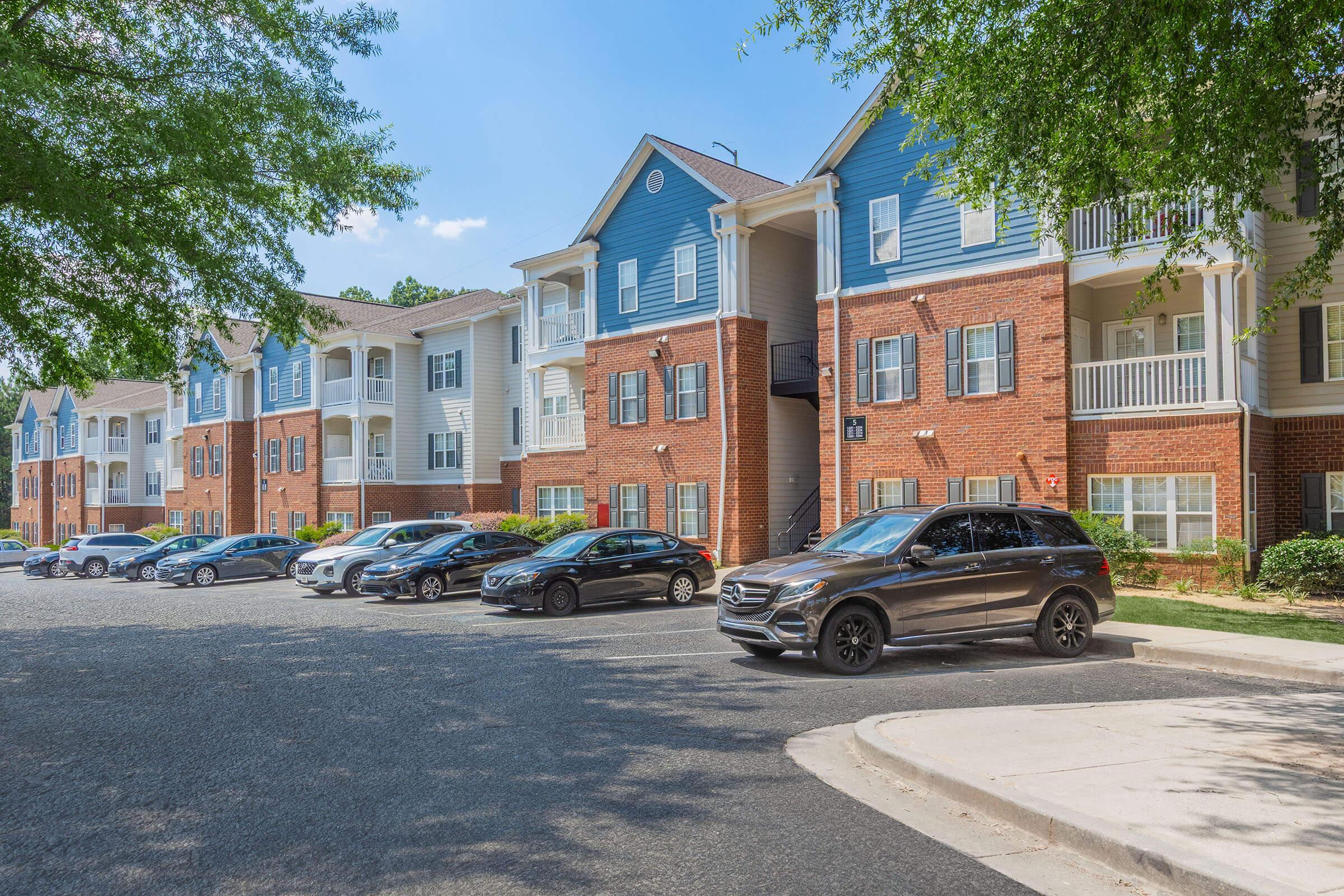 A residential apartment complex featuring several buildings in a sunny setting. The foreground includes a parking area with various cars, while the background showcases well-maintained brick and blue-sided apartments surrounded by greenery.