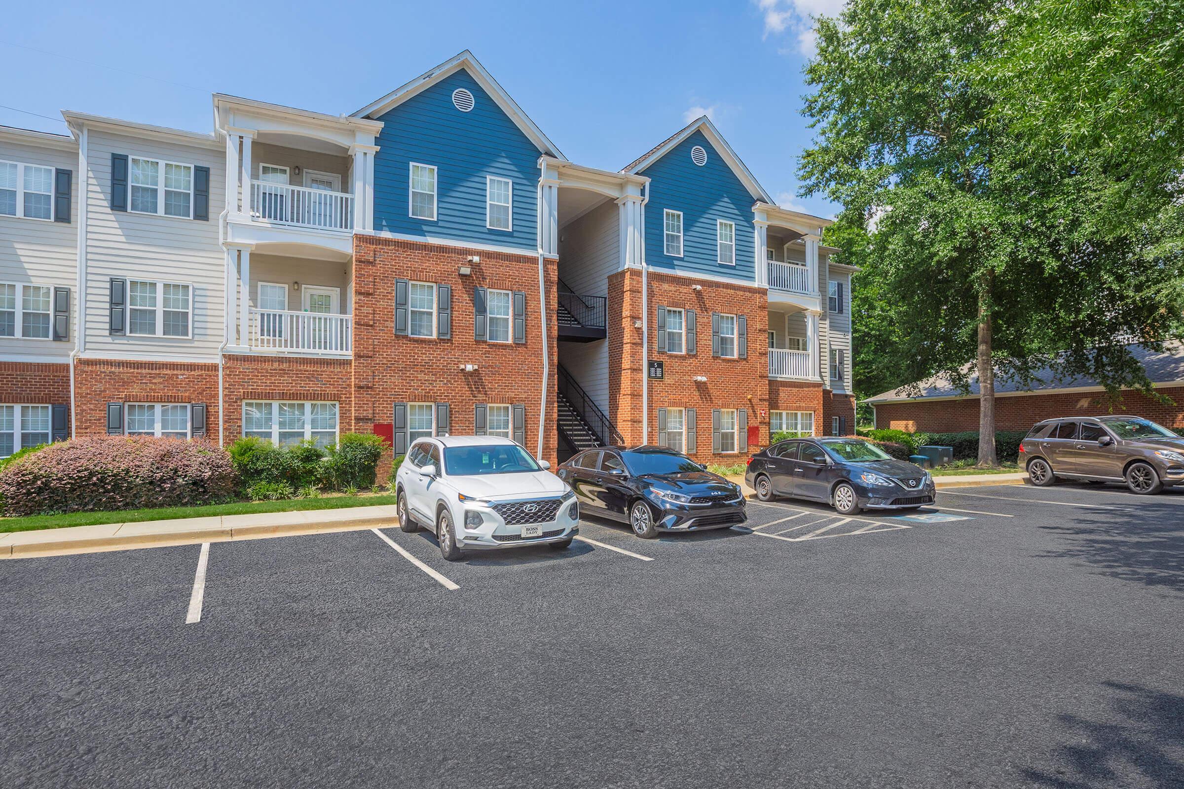 A three-story apartment building with a mix of blue and white siding, featuring balconies and stairs. In the foreground, a parking lot with several cars parked. Surrounding the building are well-maintained shrubs and green trees under a clear blue sky.