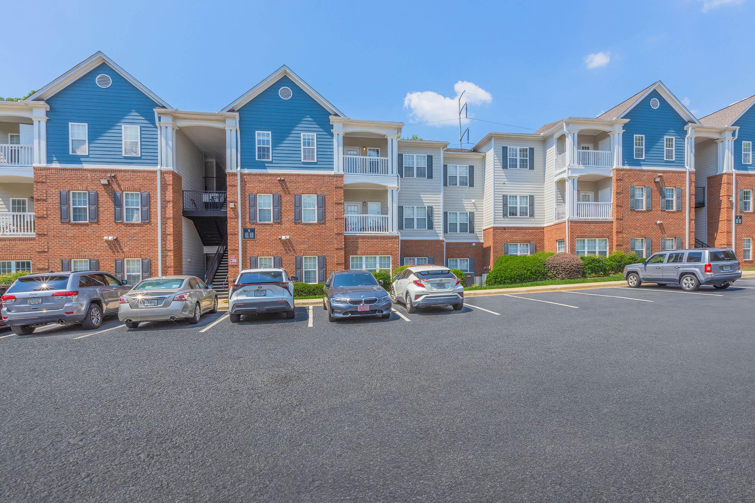 A multi-story residential building with a mix of blue and red brick exteriors. Several parked cars are visible in the foreground, along with well-maintained landscaping. The sky is clear with a few clouds, suggesting a sunny day. The overall scene depicts a suburban apartment complex.