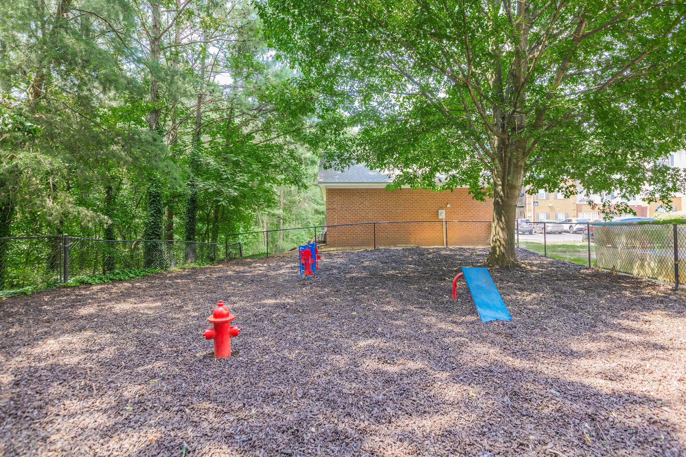 Playground area featuring a red fire hydrant, a blue slide, and a child playing near a large tree. The ground is covered with wood chips, and there is a fence surrounding the space, with a building visible in the background among greenery.