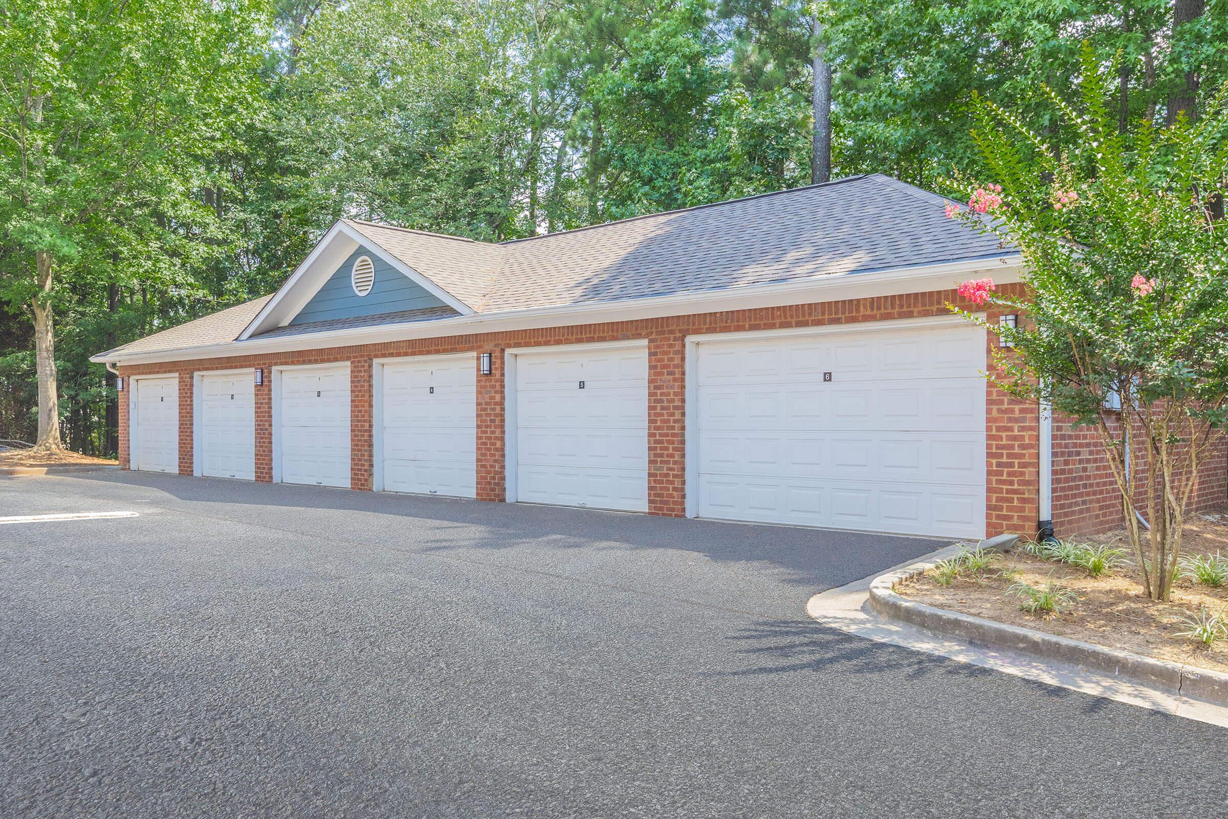 A row of six identical garage doors in a brick building, surrounded by greenery. The garages have white doors, with a sloped roof and blue accents. A paved driveway leads up to the garages, set in a peaceful outdoor environment.