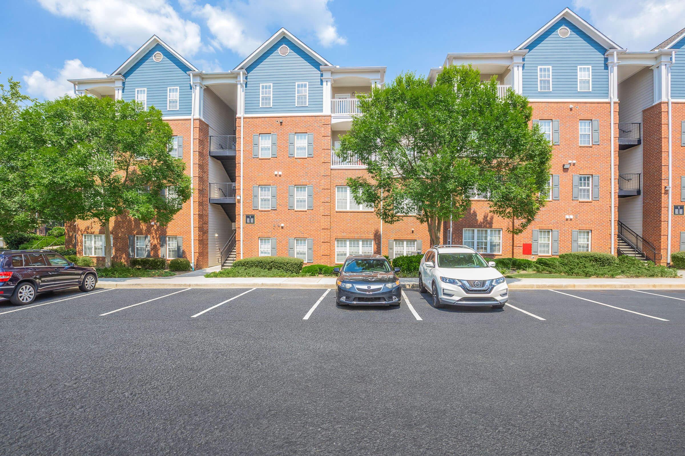 A multi-story apartment building with a mix of brick and blue siding. There are trees in front and cars parked in a lot. The building features balconies and several windows. The setting is bright and captures a clear blue sky.