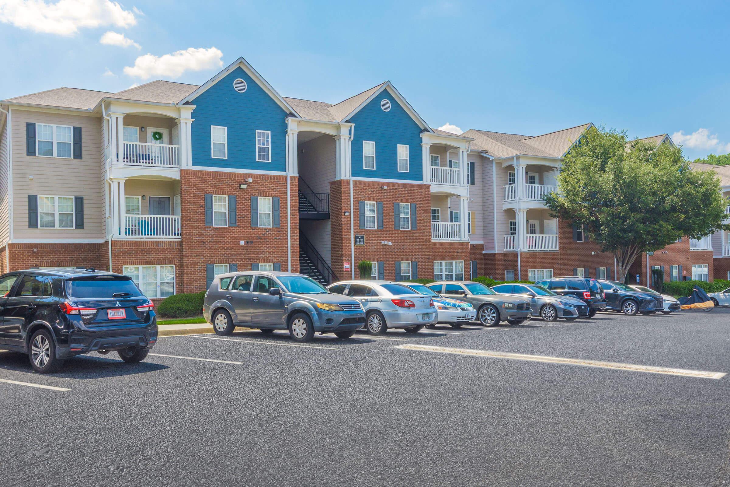 A view of a multi-story apartment building featuring a combination of blue and beige siding. The building has balconies and a staircase visible. In the foreground, there is a parking lot with several cars parked, surrounded by greenery and a clear blue sky.