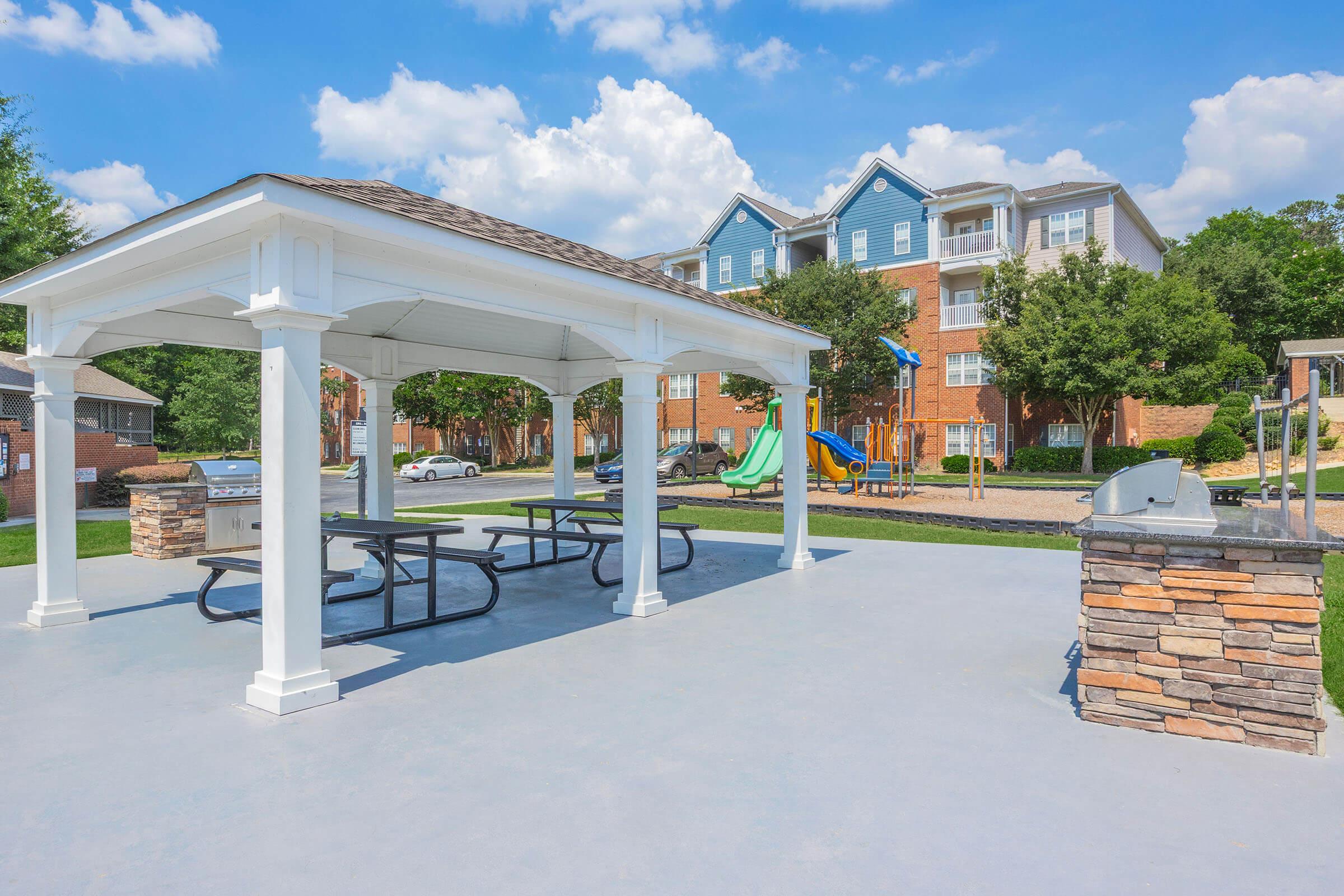 A spacious outdoor area featuring a white gazebo with picnic tables, surrounded by well-maintained green grass. In the background, there are residential buildings and a playground with slides, all under a blue sky dotted with fluffy clouds. A grill is positioned nearby for outdoor cooking.
