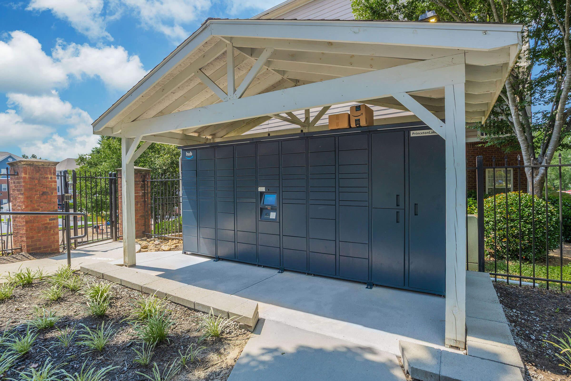 A large, dark-colored package delivery locker system located under a wooden shelter. The area is landscaped with neat plant beds, and there are residential buildings in the background. The sky is partly cloudy, indicating a clear day.