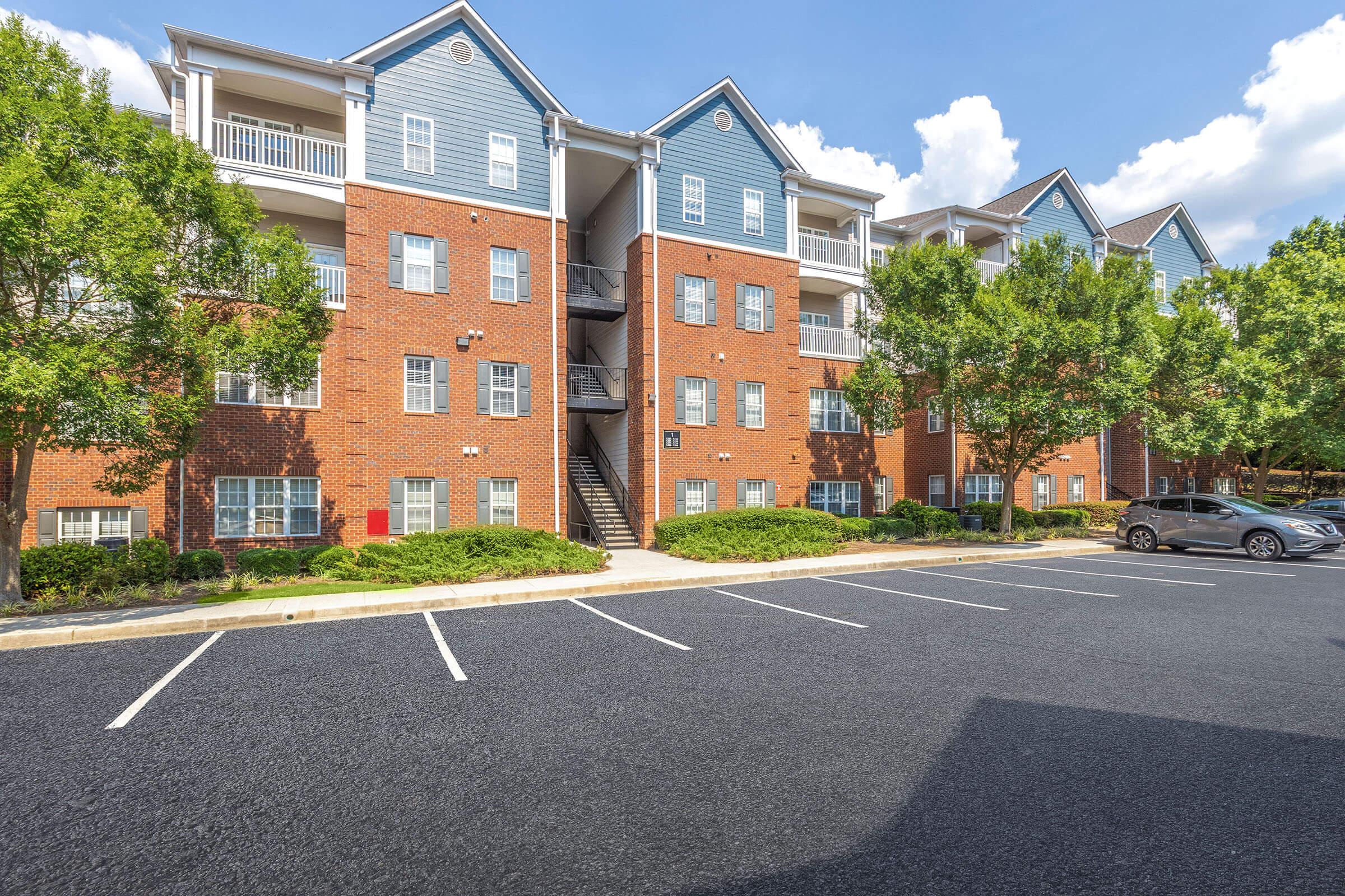 A multi-story brick apartment building with gray accents, surrounded by well-maintained shrubs and trees. There are several parking spaces in front, and the sky is clear with a few clouds. The building features balconies and stairwells for access to different floors.