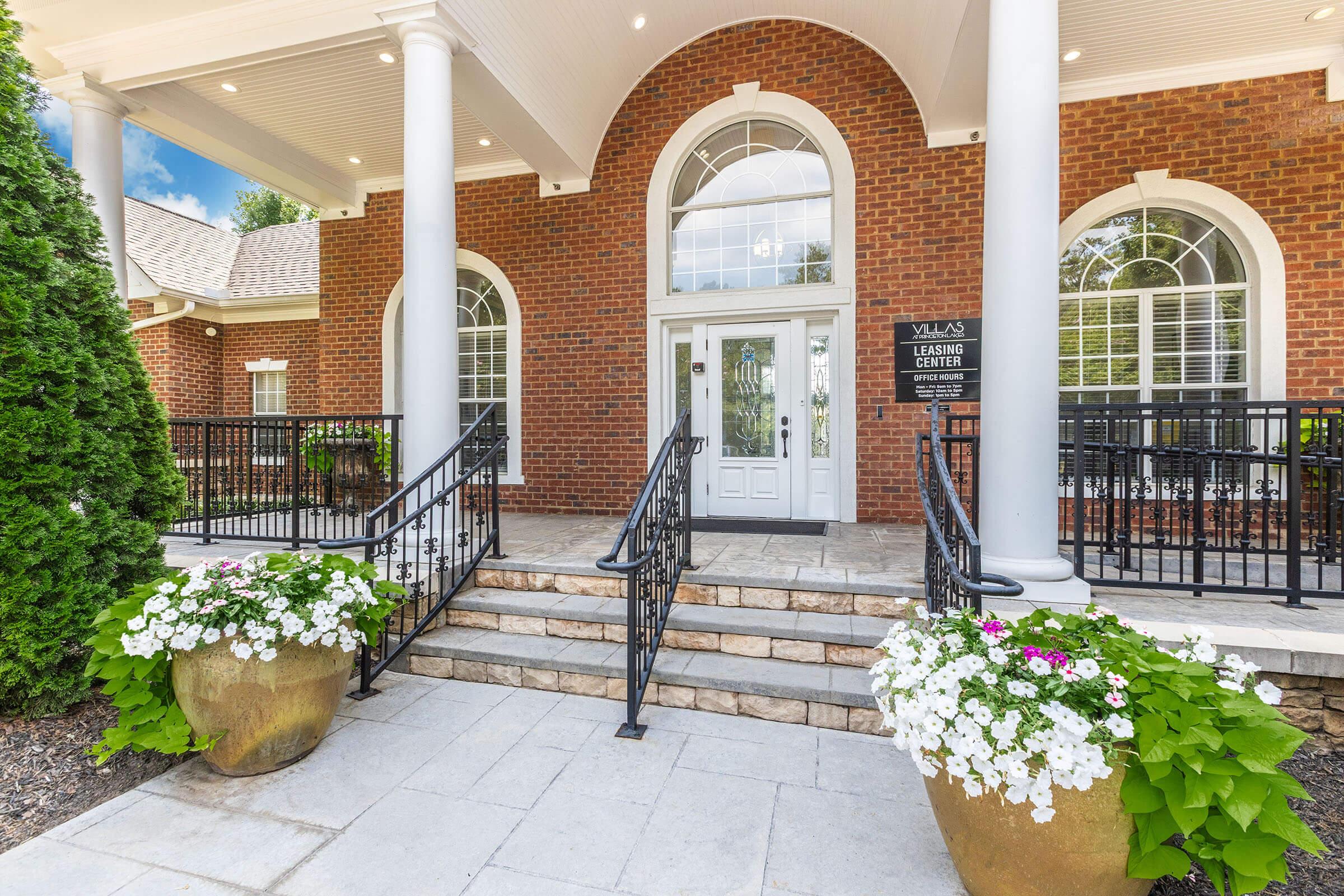 A welcoming entrance to a leasing center featuring large brick columns, an elegant door, and bright white windows. The steps are flanked by large planters with colorful flowers, adding to the inviting atmosphere of the property.