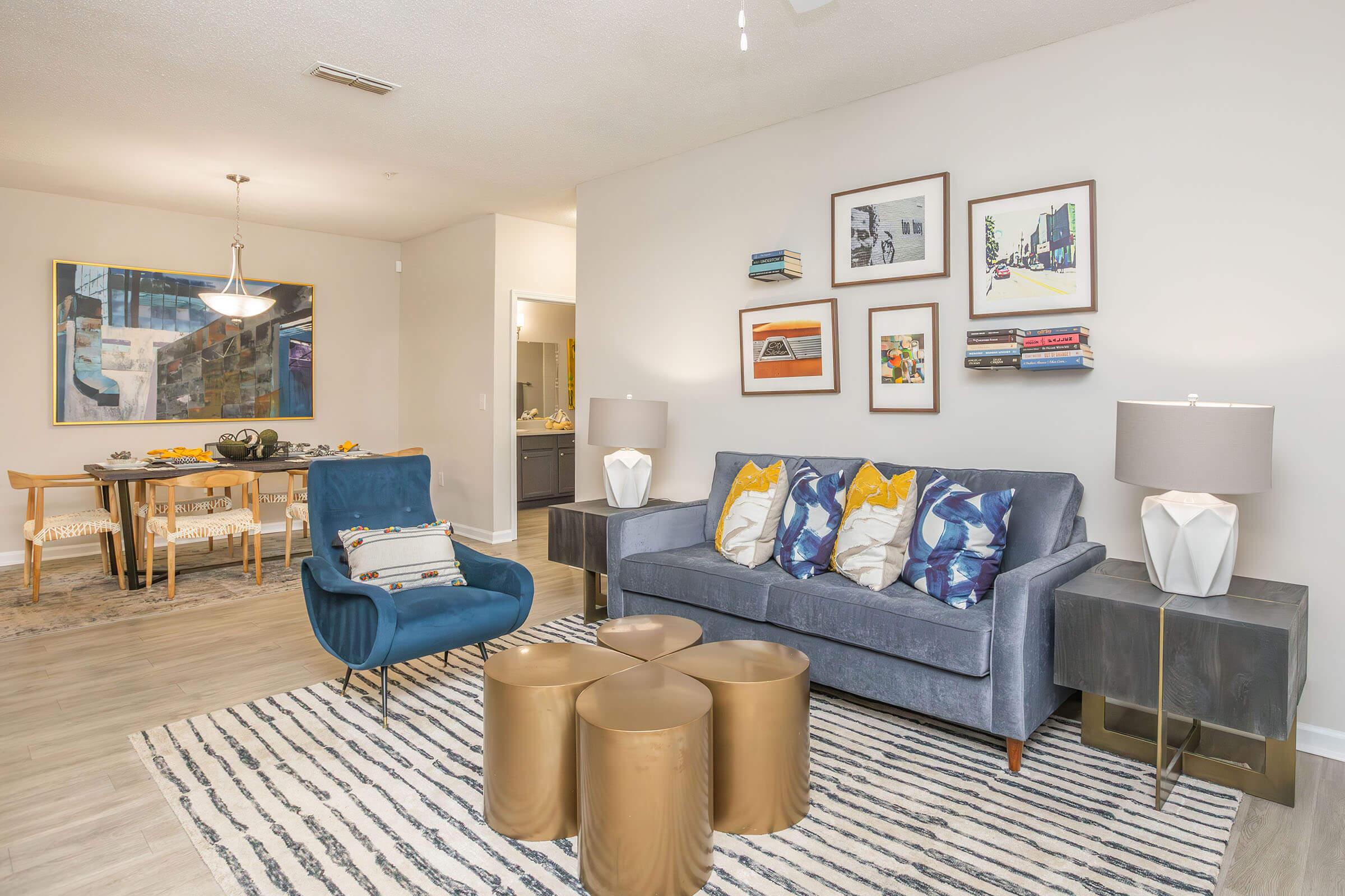 A stylish living room featuring a blue velvet chair, a gray sofa with colorful pillows, and gold accent tables. The walls are decorated with framed artwork. A dining area is visible in the background, with a modern table and chairs, and a pendant light above. The flooring is light-colored wood, adding to the contemporary feel.
