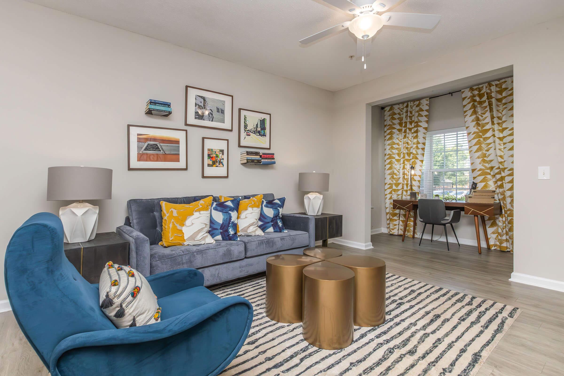 A modern living room featuring a blue sofa adorned with yellow and blue pillows, circular gold coffee tables, and stylish decor. On the wall, there are framed artworks. A cozy workspace with a wooden desk and patterned curtains is visible in the background. The space has a light, airy vibe with wooden flooring.