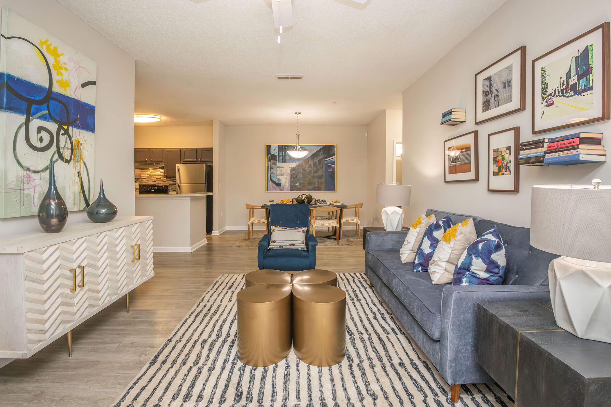 A stylish living room featuring a blue sofa with colorful throw pillows, two gold coffee tables, and decorative vases. The walls are adorned with framed artwork and books. A dining area with a wooden table and chairs is visible in the background, along with a modern kitchen. Bright, neutral tones create an inviting atmosphere.