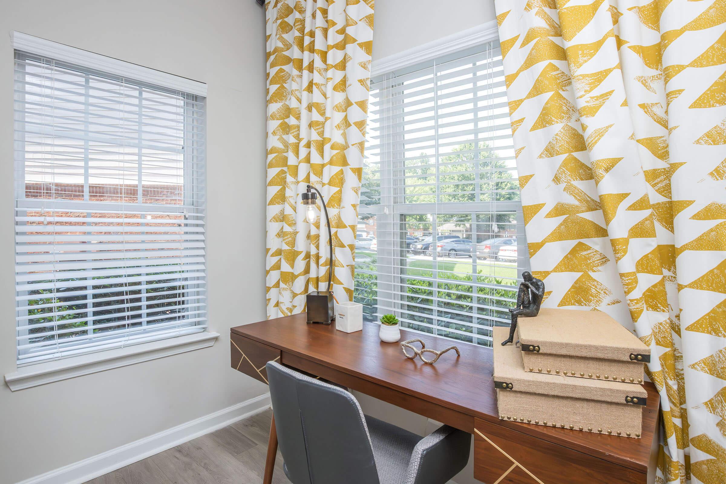 A well-lit home office corner featuring a wooden desk with decorative boxes, a small plant, and glasses. Two large windows with white blinds are dressed with yellow and white patterned curtains, bringing a cheerful ambiance to the space.