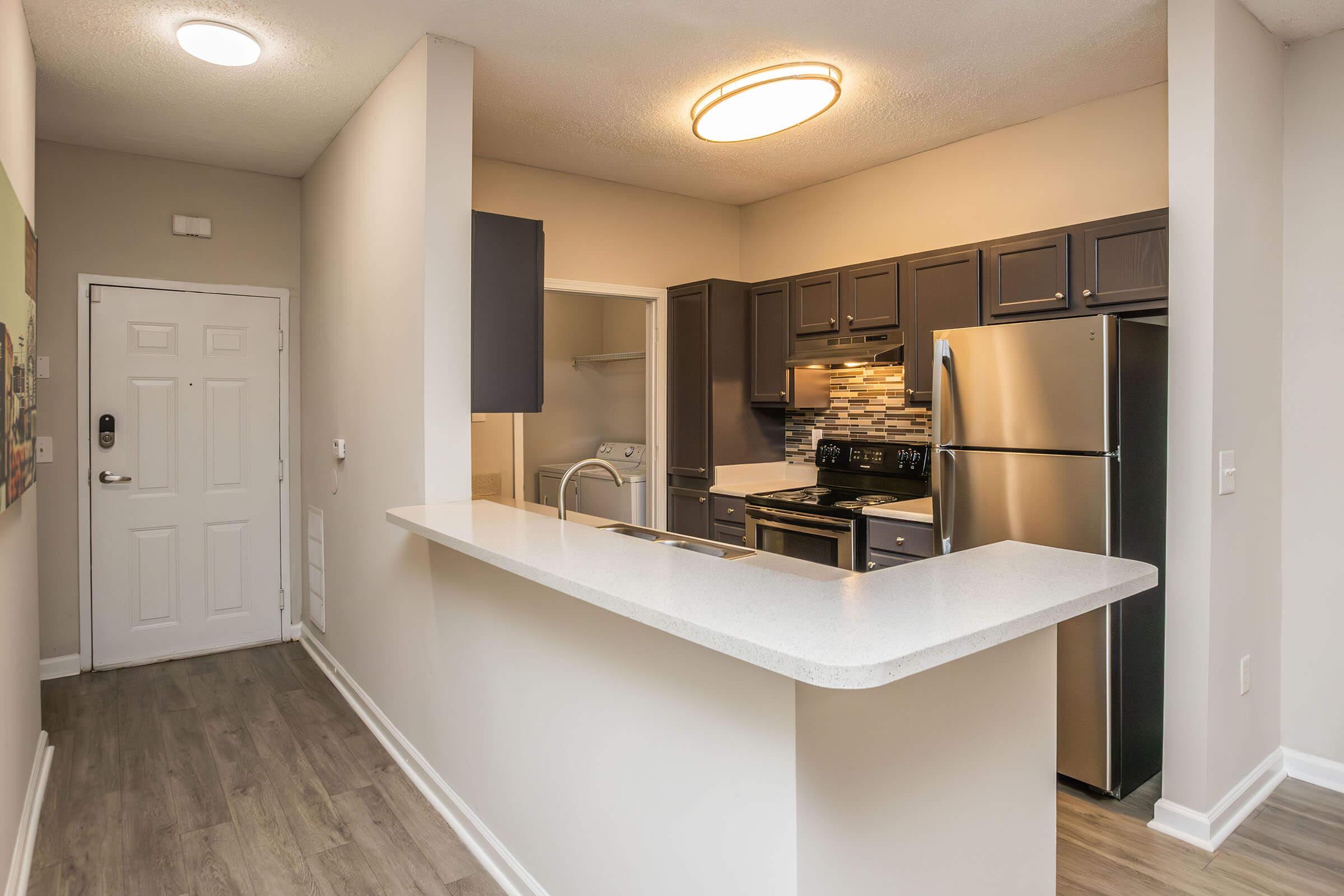 Modern kitchen and living area featuring a bar counter, stainless steel appliances, and dark cabinetry. The open floor plan includes a front door, small closet, and a light fixture. Elegant design with a neutral color palette and wood flooring.