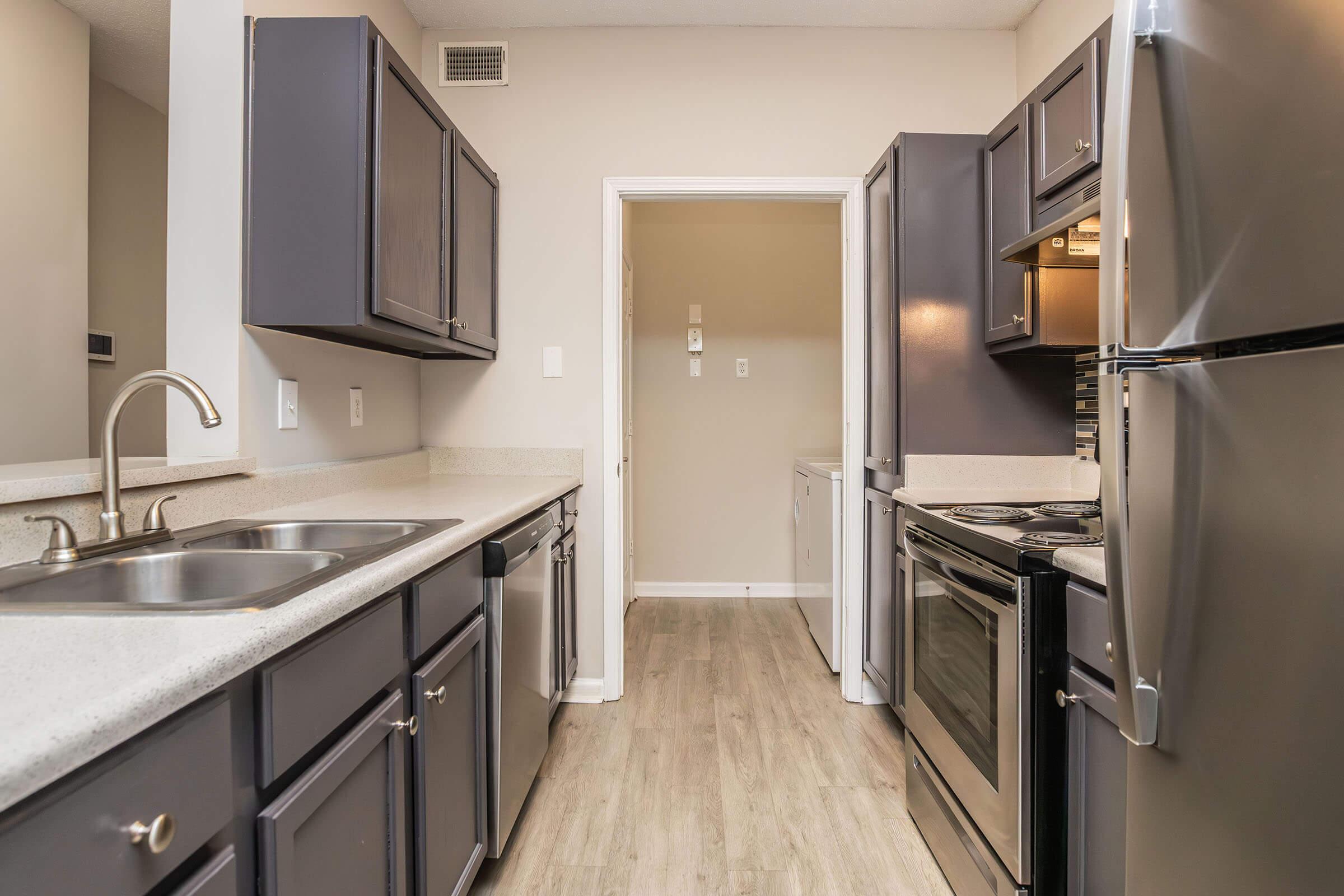 Modern kitchen featuring dark cabinetry, stainless steel appliances, a double sink, and a smooth countertop. The layout includes an oven, microwave, and a view into a laundry area through an open doorway. The floor is light-colored laminate, enhancing the spacious feel of the room.