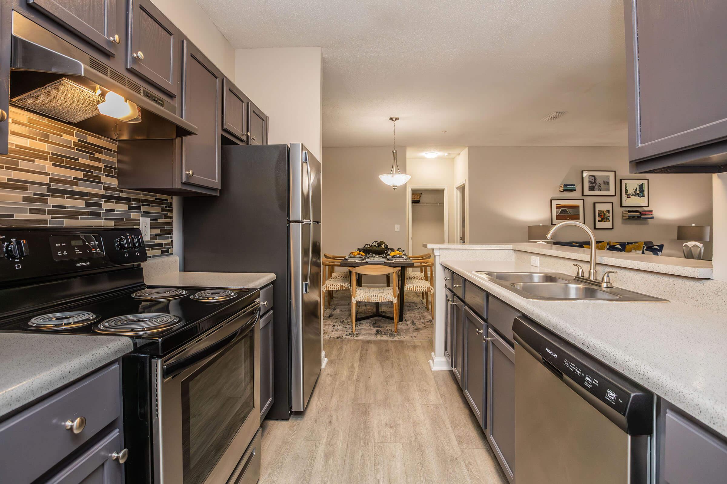 Modern kitchen featuring dark cabinetry, stainless steel appliances, and a stone backsplash. Includes a double sink and an electric stove with four burners. A dining area is visible in the background, furnished with a table and chairs, plus decorative wall art.