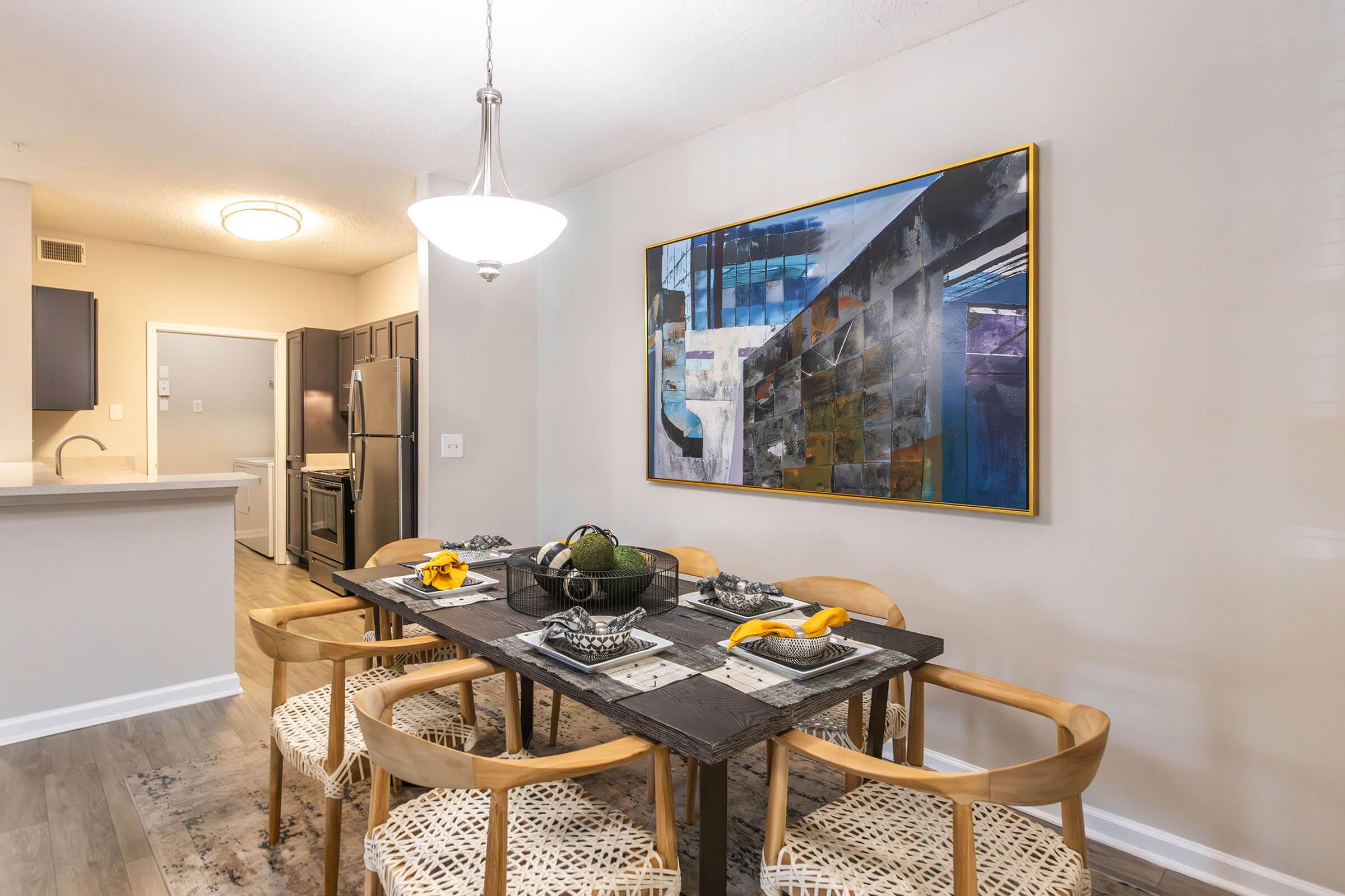 A modern dining area featuring a wooden table surrounded by four stylish chairs. The table is set with decorative items and a fruit bowl. Above hangs a contemporary light fixture. A large abstract painting adorns the wall, and a kitchen area is visible in the background with dark cabinets and stainless steel appliances.