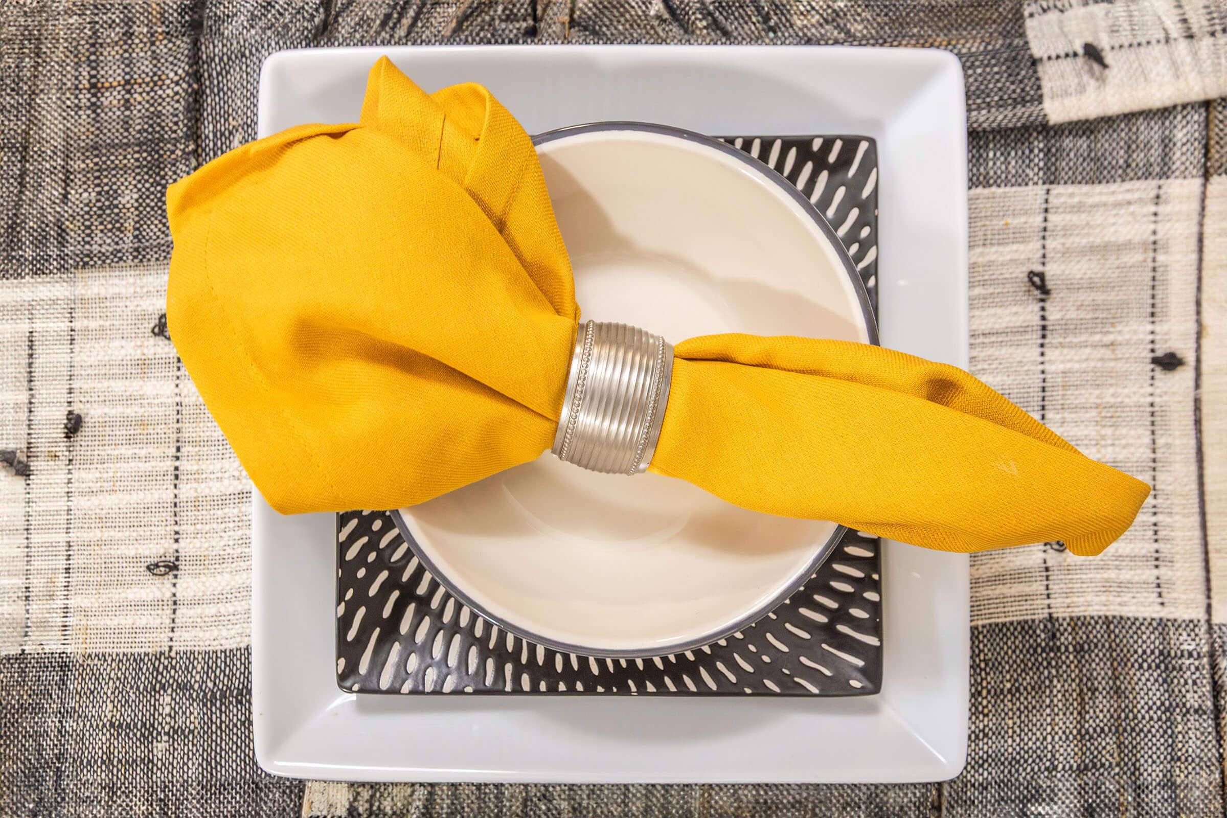 A neatly arranged dinner setting featuring a white plate on a decorative black and white charger. A vibrant yellow napkin is elegantly folded and held with a silver napkin ring, adding a pop of color to the table setting. The background shows a patterned tablecloth.