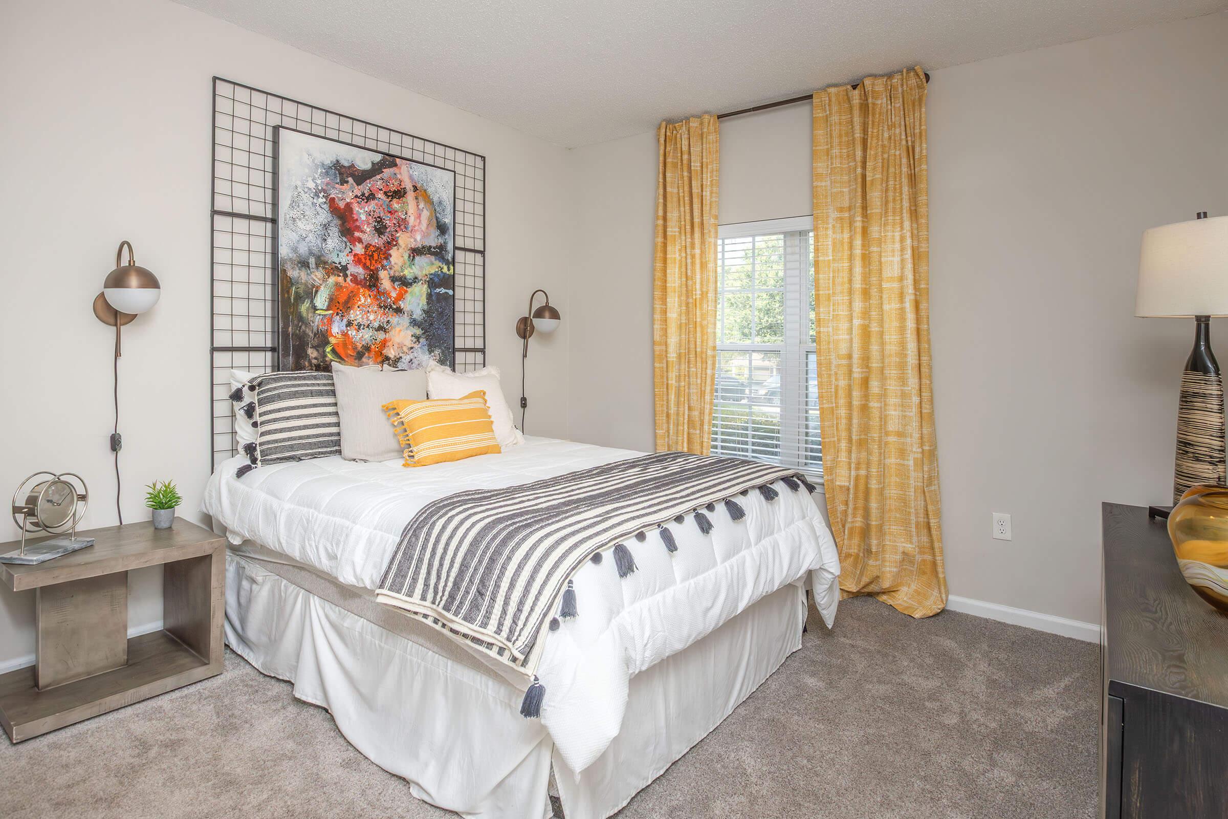 A well-decorated bedroom featuring a queen-sized bed with a white comforter and decorative pillows. The walls are adorned with a colorful abstract painting and there are light-colored curtains at the window. A bedside table with a lamp and a small plant complement the cozy atmosphere.