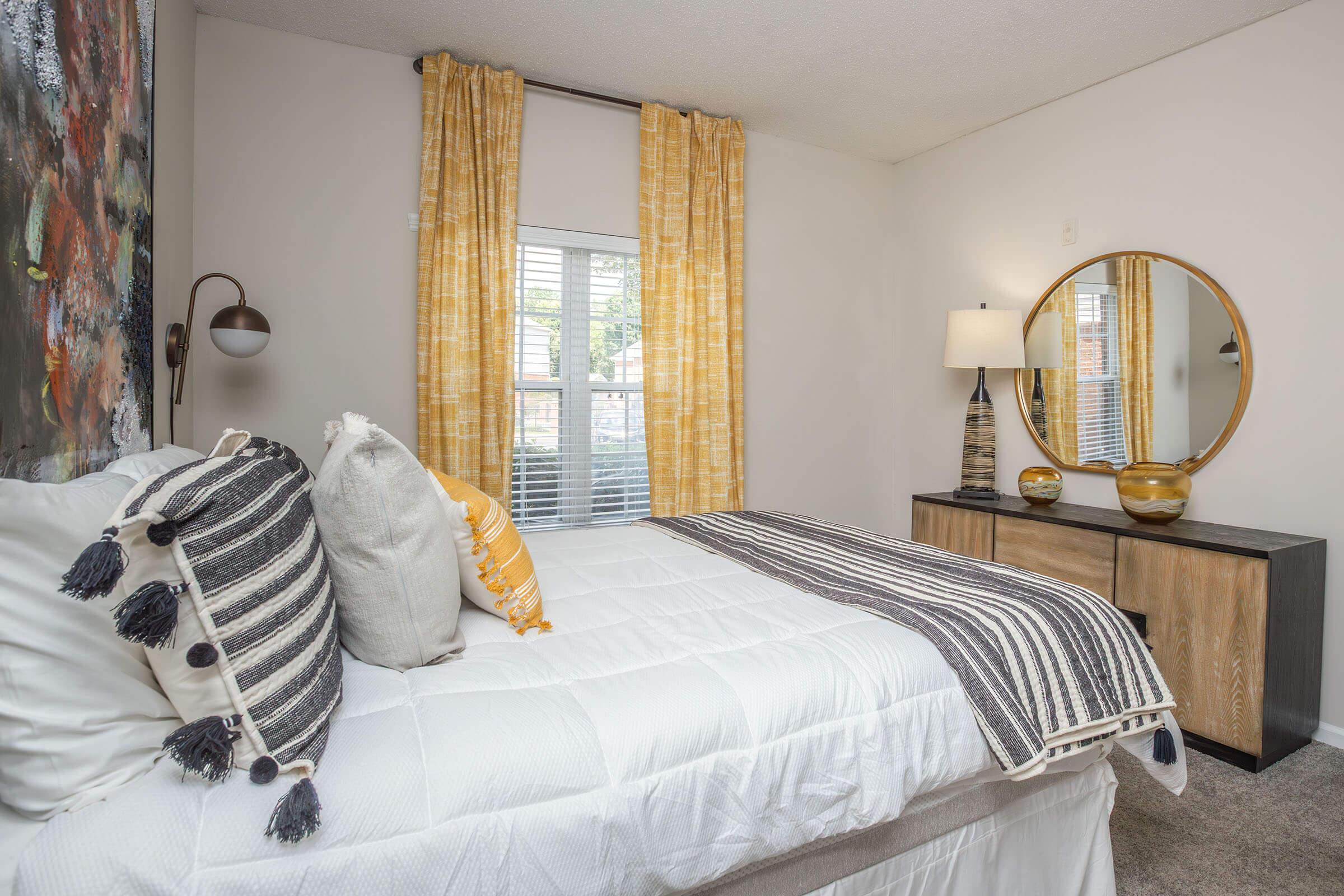 A cozy bedroom featuring a neatly made bed with decorative pillows, a round mirror above a modern dresser, and warm yellow curtains framing a window. The walls are adorned with artwork, and the overall decor combines comfort with contemporary style.
