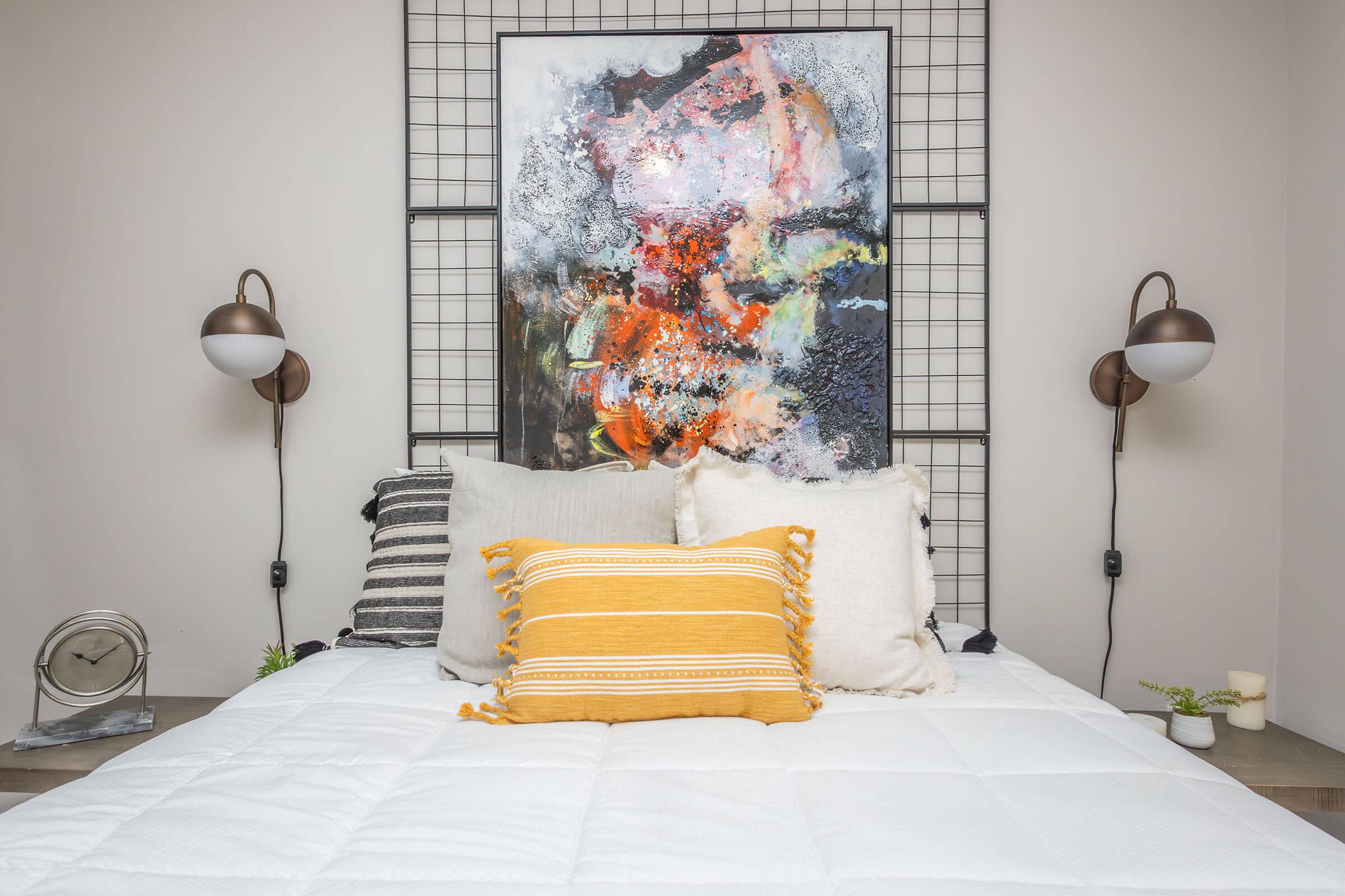 A cozy bedroom setting featuring a white bed with multiple decorative pillows, including a yellow-striped one. Above the bed, there is a large, vibrant abstract painting. On either side of the bed, there are stylish wall-mounted lamps. Small plants and a clock are visible on the bedside table.
