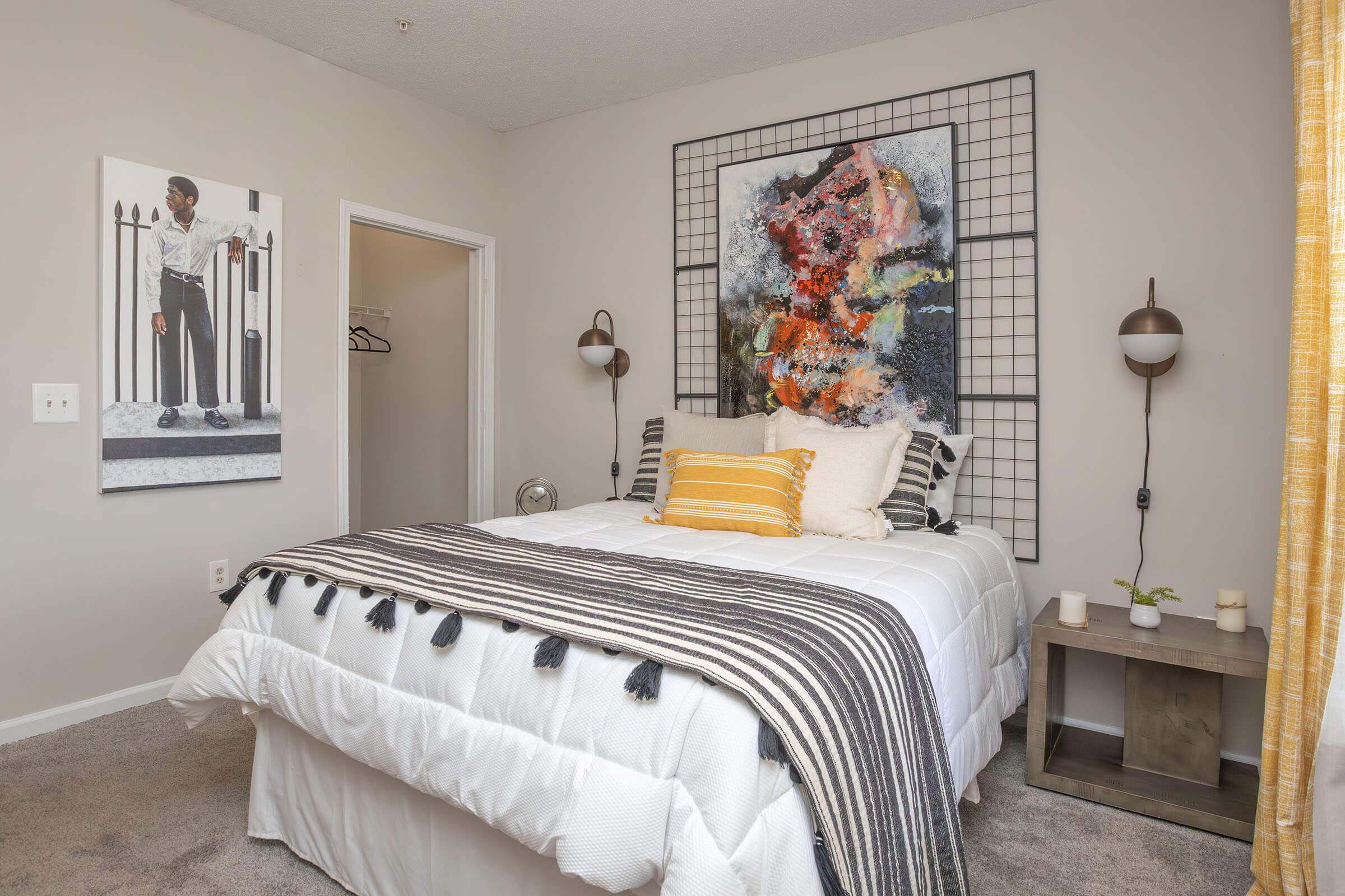 A cozy bedroom featuring a neatly made bed with a striped blanket and decorative pillows. There's a large, colorful abstract painting above the bed and a stylish wall art photo of a person in contemporary attire. The room has soft lighting and a warm color scheme, with a wooden nightstand beside the bed.
