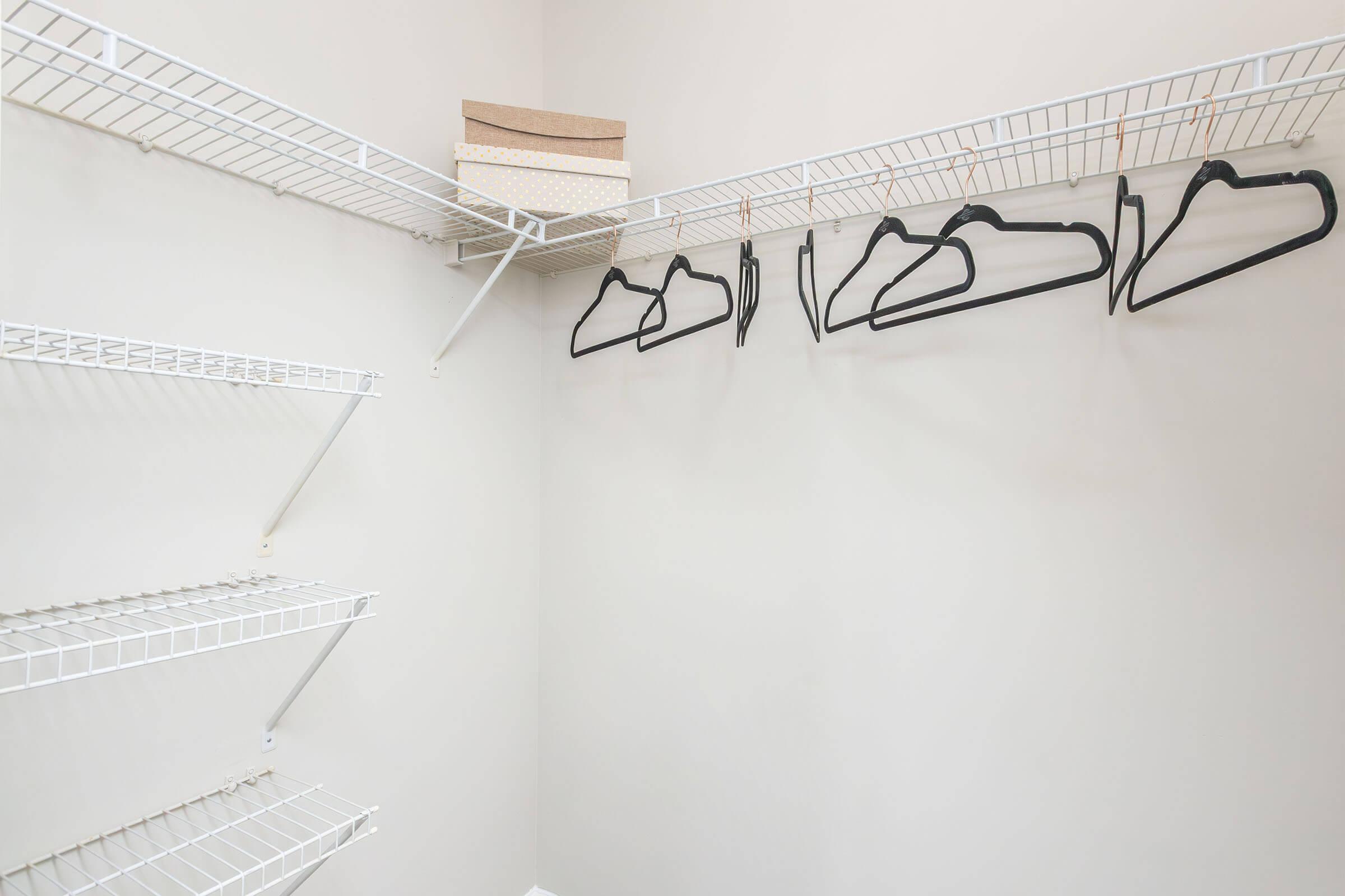 A neatly organized closet featuring white wire shelves. Several black hangers are hanging on one shelf, and two decorative boxes are placed on the top shelf. The walls are a light neutral color, creating a clean and spacious appearance.