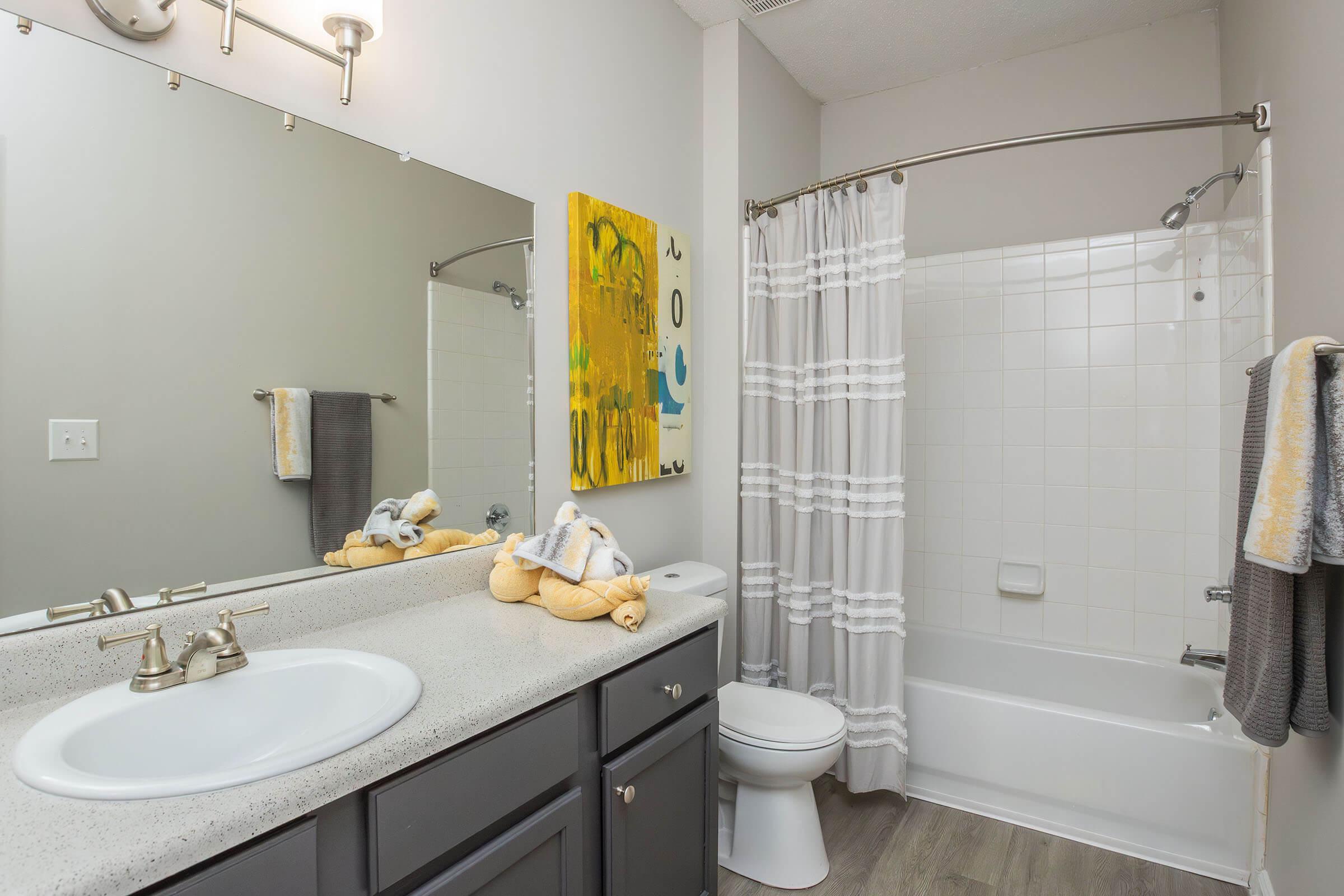 A well-lit bathroom featuring a double sink vanity with a large mirror, decorative yellow wall art, a bathtub with a shower curtain, and neatly arranged towels. The color scheme includes shades of gray, yellow, and white, creating a modern and clean look.