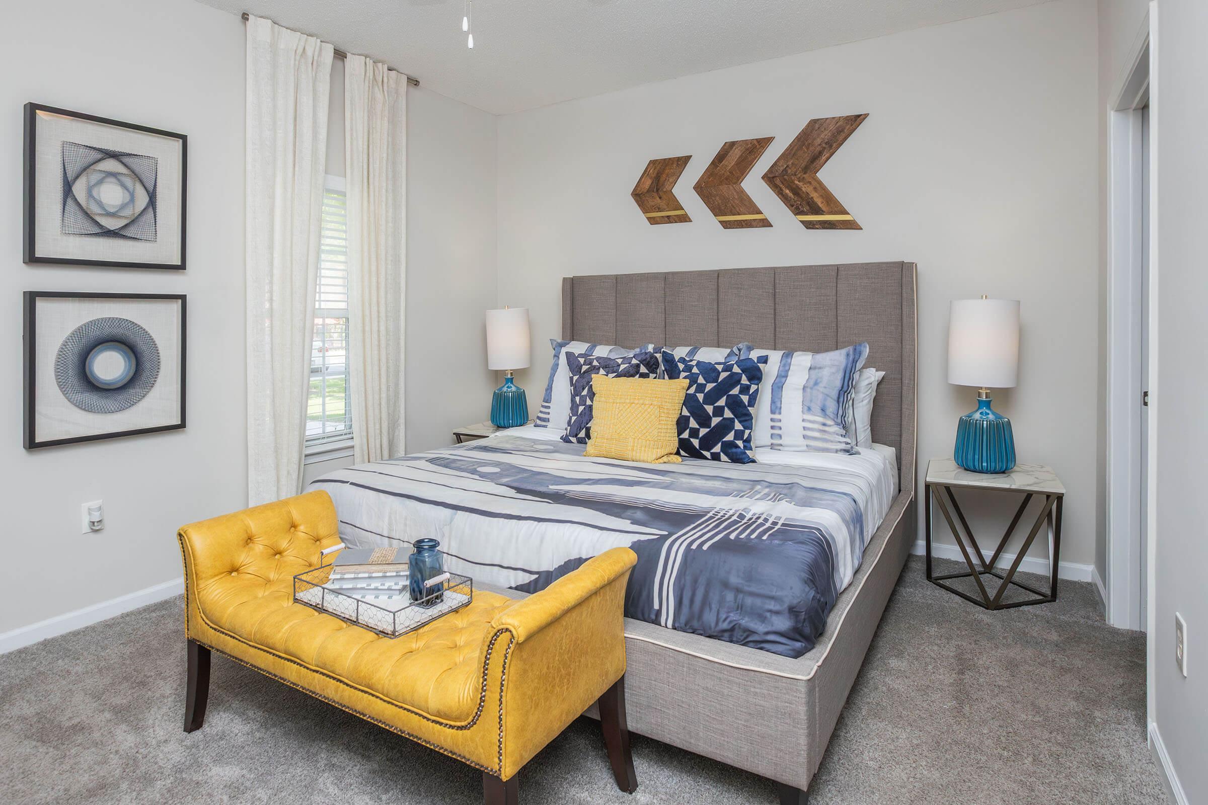 Modern bedroom featuring a gray upholstered bed with patterned bedding, a yellow tufted bench at the foot, and two bedside tables with blue lamps. Decor includes framed geometric art on the walls and wooden arrow accents above the bed. The space has light-colored curtains and gray carpet.