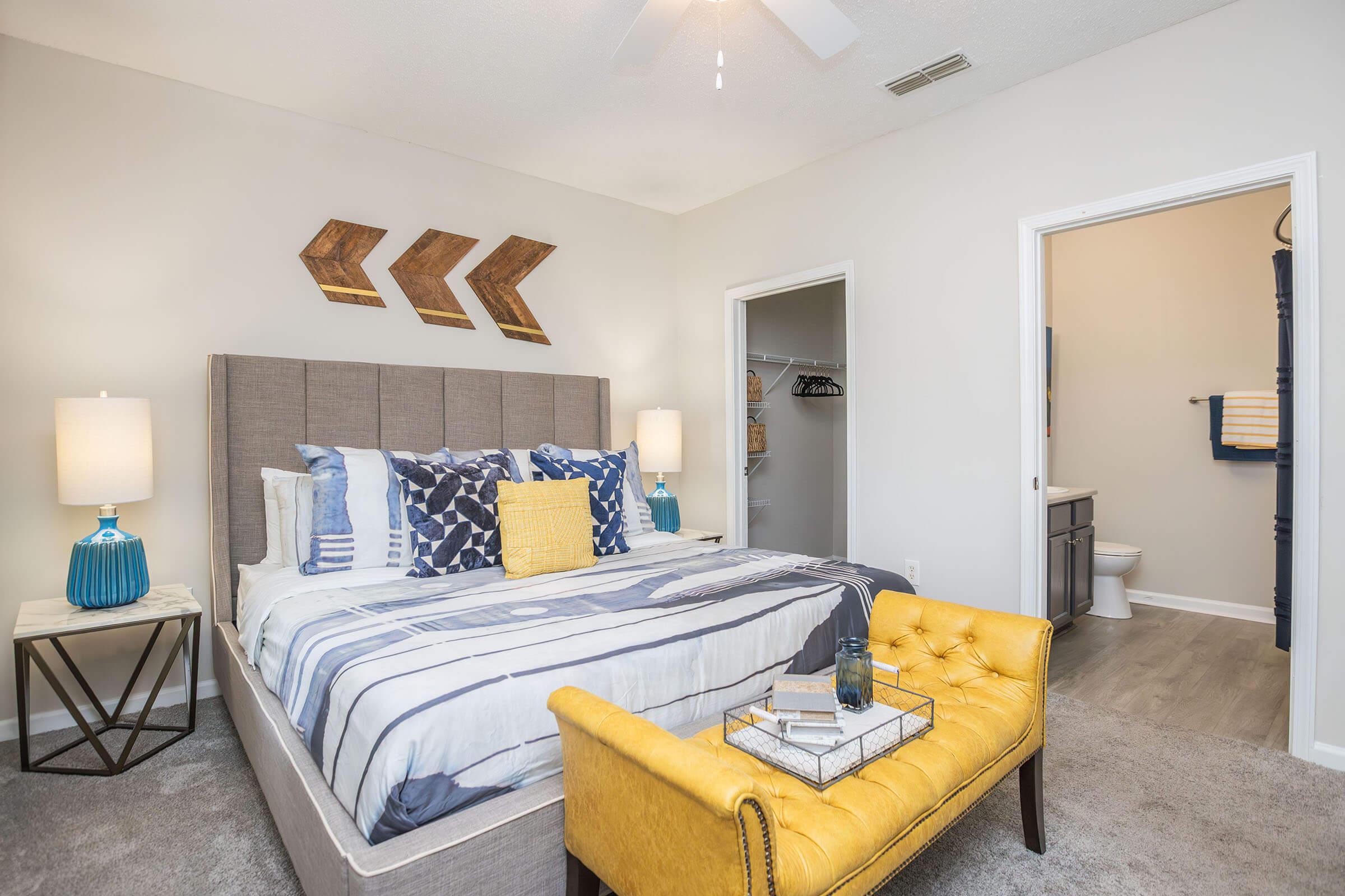 A stylish bedroom featuring a neatly made bed with decorative pillows, a yellow tufted bench at the foot, bedside tables with lamps, and modern wall decor. A bathroom entry is visible in the background, showcasing a contemporary design. The room has light-colored walls and a fan overhead.