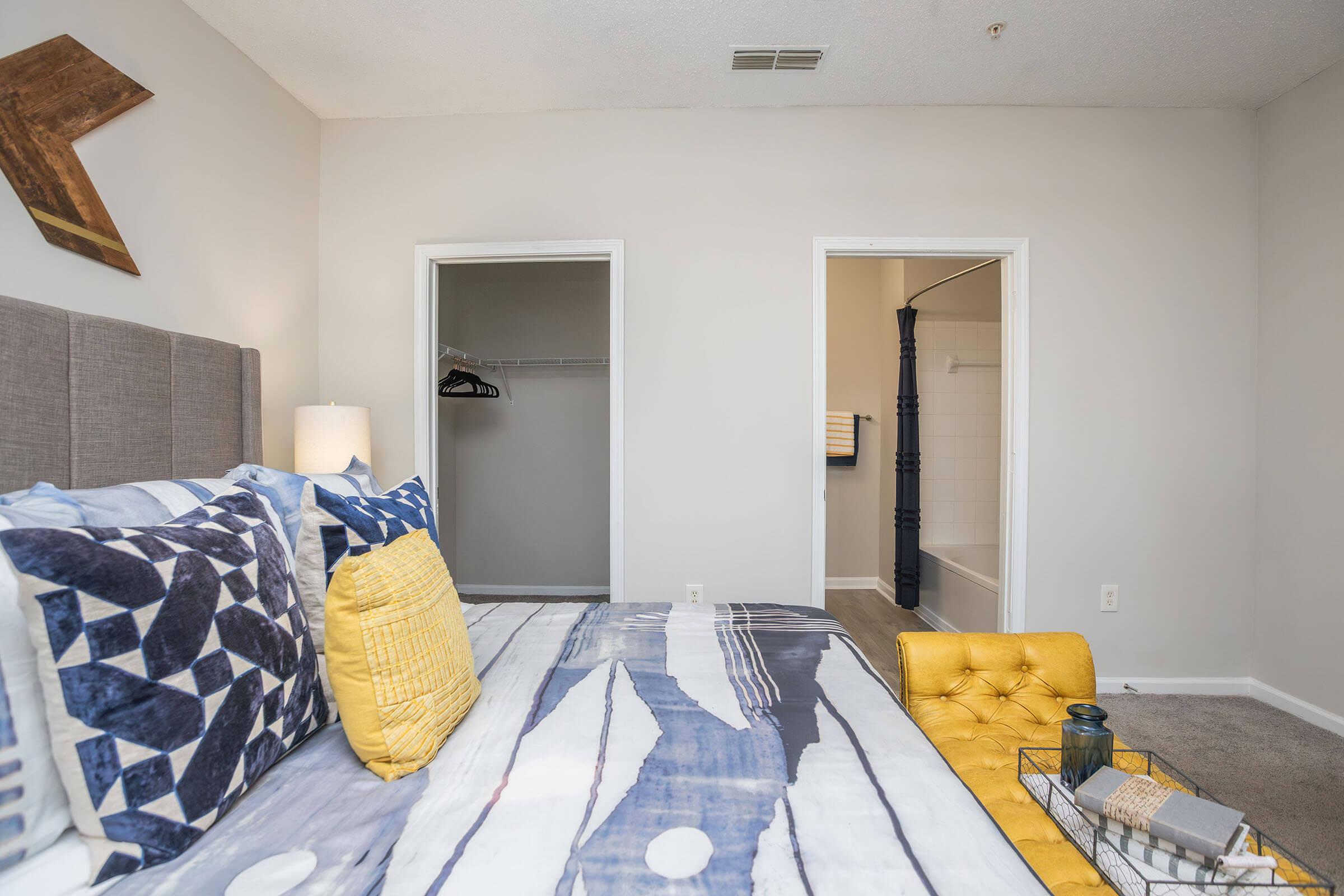 A modern bedroom featuring a gray upholstered bed with decorative pillows, a yellow accent pillow, and a cozy throw blanket. A wardrobe is visible in an adjacent room, along with a bathroom entrance. The walls are painted light colors, and the floor is carpeted, creating a warm and inviting atmosphere.