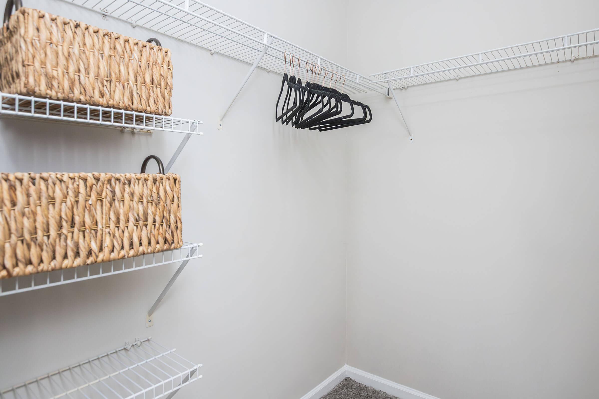 A spacious, empty closet featuring white wire shelving. Two shelves are filled with woven storage baskets, while the wall has several black hangers neatly arranged. The room has a light, neutral color scheme and a carpeted floor.